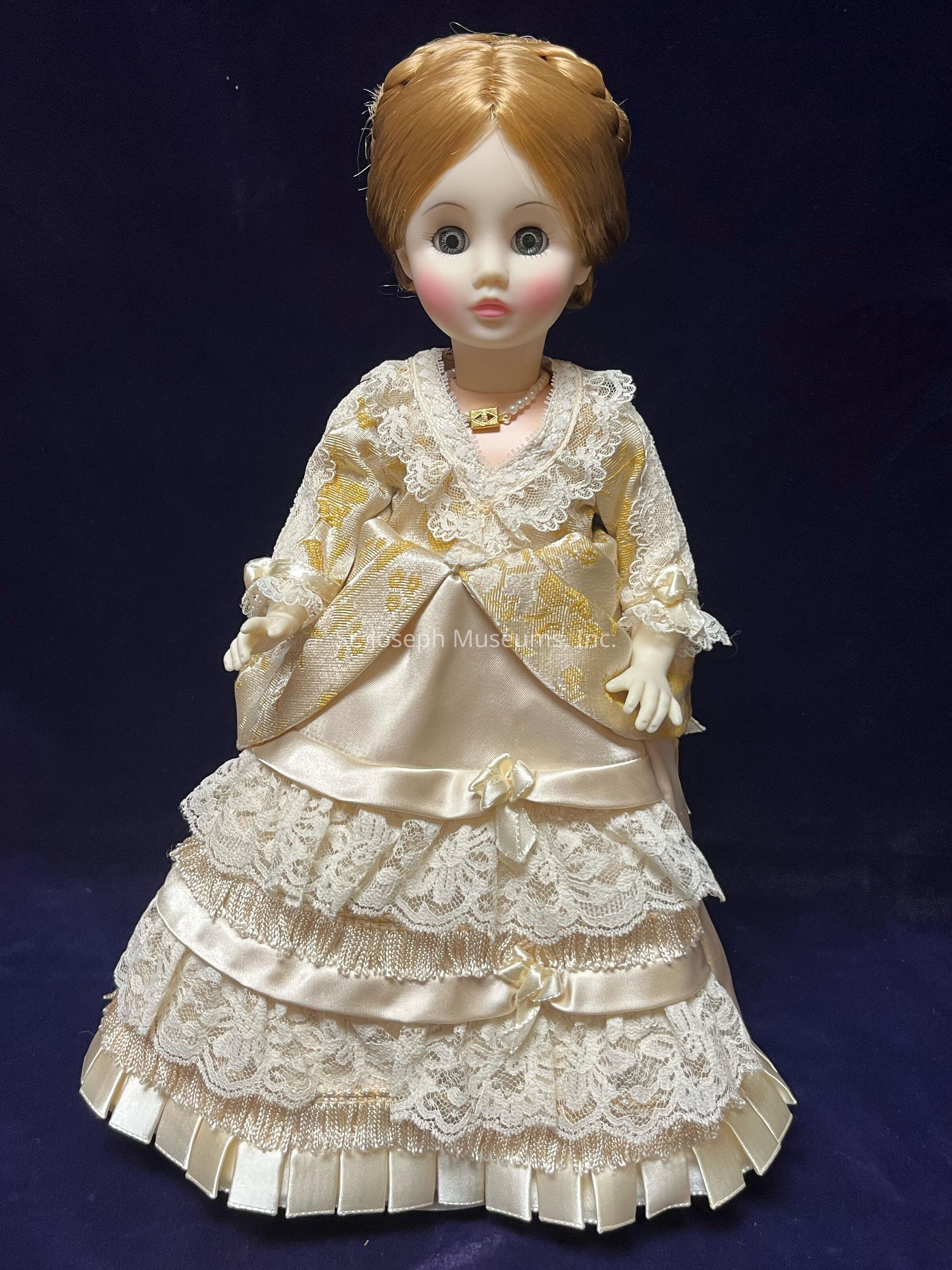 The image features a Madame Alexander doll in the likeness of Lucy Webb Hayes. Her hair is styled in an elegant updo and the doll is dressed in a cream-colored gown and adorned with intricate lace detailing and satin ribbons. The bodice of the dress is embellished with lace and a small gold-colored medallion. The doll's layered skirt is accentuated with delicate lace patterns and satin trim.