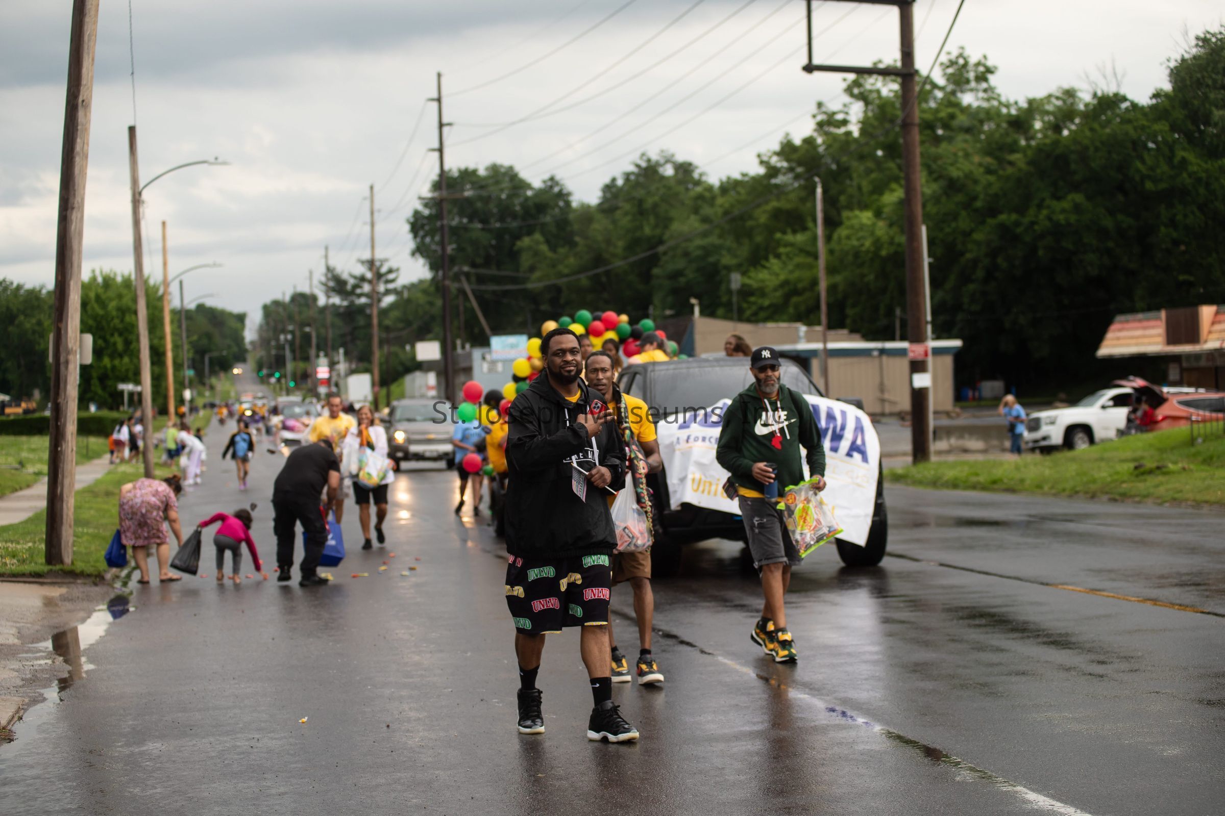 The image depicts a lively street scene during a parade, where a group of people are walking on a wet road, suggesting recent rain. In the foreground, two men are prominently featured, one holding a bag filled with candy, while another carries colorful balloons, creating a festive atmosphere. Behind them, a vehicle adorned with a banner and more balloons moves slowly, with additional participants following along. On either side of the road, people are bent over, picking items from the ground, possibly candy thrown during the parade. Trees and utility poles line the road, adding to the suburban backdrop, while the cloudy sky suggests an overcast day.