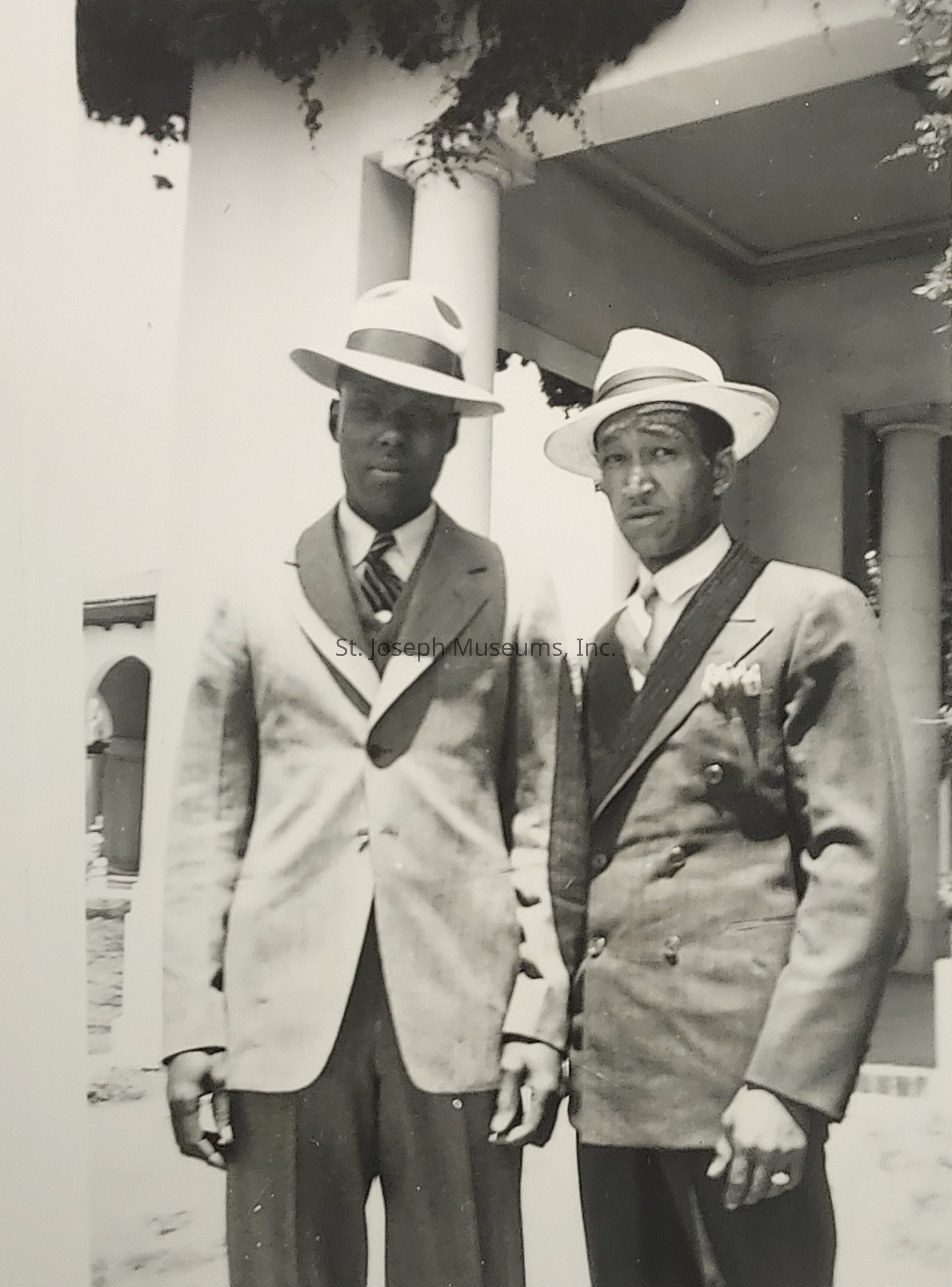 The image shows two men standing side by side, dressed in elegant suits with ties and vests, and wearing stylish hats typical of early to mid-20th century fashion. The man on the left wears a lighter-colored suit, while the man on the right is in a darker suit adorned with a boutonnière, indicating perhaps a formal event or gathering. Both have serious expressions, and their attire suggests attention to detail and a sense of sophistication. They stand in front of a building with white pillars and an arched entrance, partly obscured by foliage, adding a touch of classic architectural elegance to the scene.