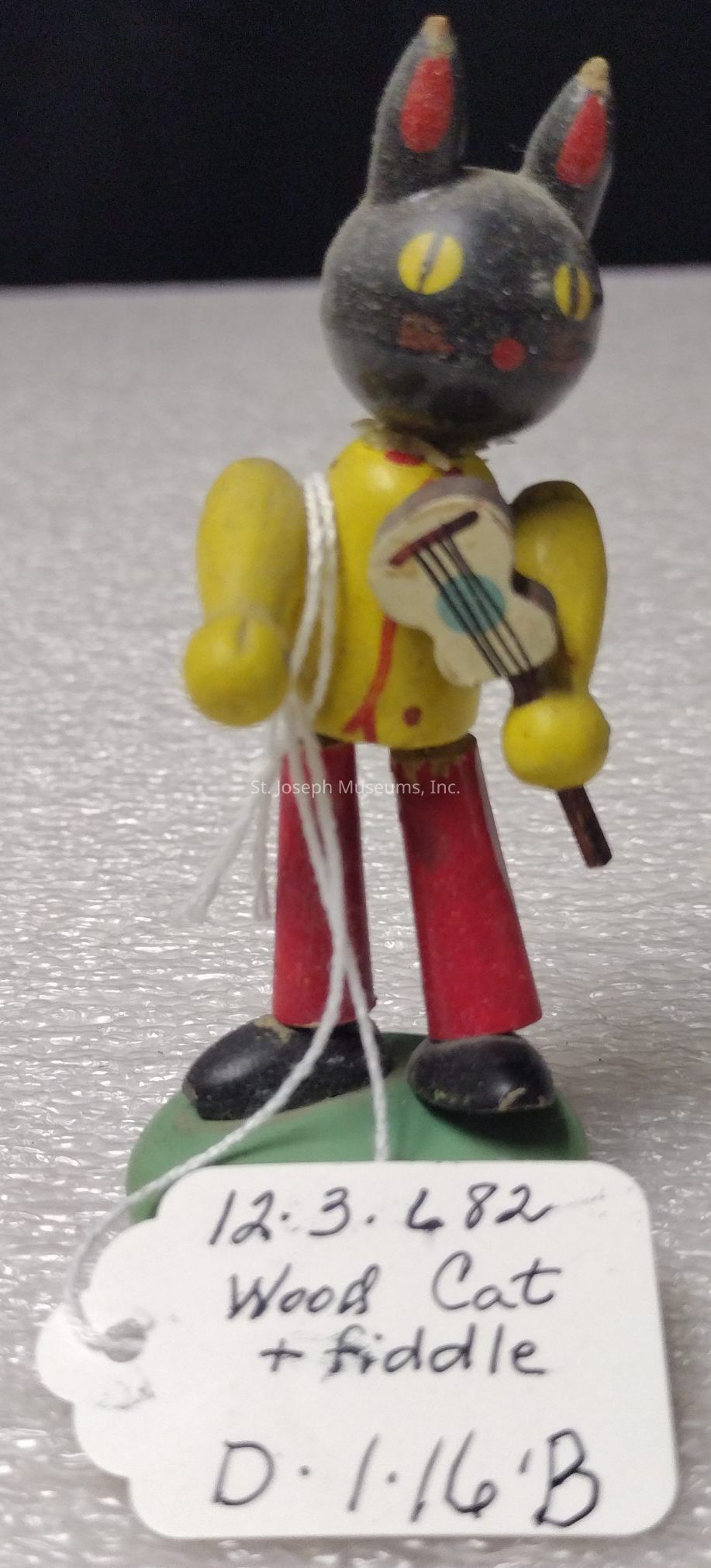 The image depicts a small wooden figurine of a cat standing upright, featuring a black head with pointed ears and yellow eyes, while wearing a yellow shirt and red pants. The cat is holding a fiddle to one side, and there is a white string attached to it. The figurine stands on a green base, and in front, there's a white paper tag with handwriting that reads, "12.3.482 Wood Cat + Fiddle D.1.16.B." The overall appearance of the figurine suggests a playful, whimsical design, characteristic of folk art. The surface on which it stands appears to be a textured, shiny material.