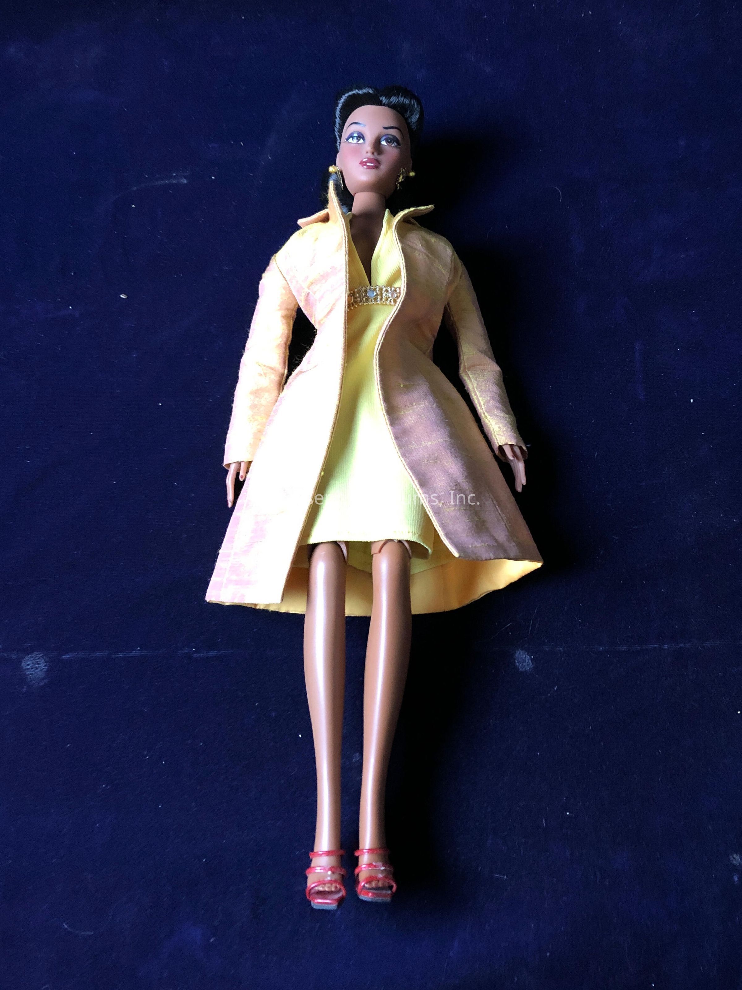 The image depicts a fashion doll lying on a dark blue, perhaps velvet, fabric surface. The doll wears a stylish outfit consisting of a bright yellow dress, which is slightly sparkly, accented with a decorative feature at the neckline that resembles a cluster of little jewels or sequins. Over the dress, the doll wears a tailored, light yellow coat with a nice sheen, seemingly made from a textured material. Her expression is poised, with a gentle smile and striking facial features including prominent eyes and softly painted lips. Her hair is styled neatly, likely in an updo or another elegant fashion. Adorning her feet are red strappy high heels that add a pop of color against her otherwise yellow-themed attire. The background, which serves as a simple and plain contrast, highlights the vibrant colors and elegant details of the doll's ensemble. Overall, the doll exudes a sense of style and sophistication.
