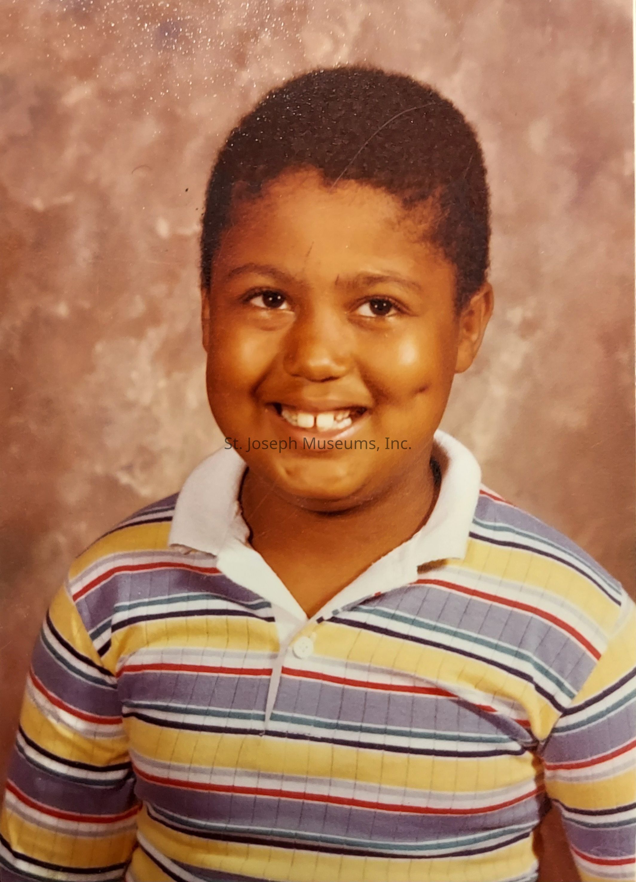The image depicts a young child with a joyful smile, showing their front teeth. The child is wearing a colorful, striped polo shirt with shades of yellow, blue, red, and white. The background is a textured, neutral-colored backdrop, suggesting that this photograph might have been taken in a studio setting, possibly as a school picture or a portrait session. The child's eyes are bright and expressive, conveying a sense of innocence and happiness. The hair is short and neatly styled, complementing the overall cheerful and clean appearance. The lighting is soft, highlighting the child's warm skin tone and the natural shine on their face. The image evokes a sense of nostalgia and captures a moment of youthful exuberance and simplicity.
