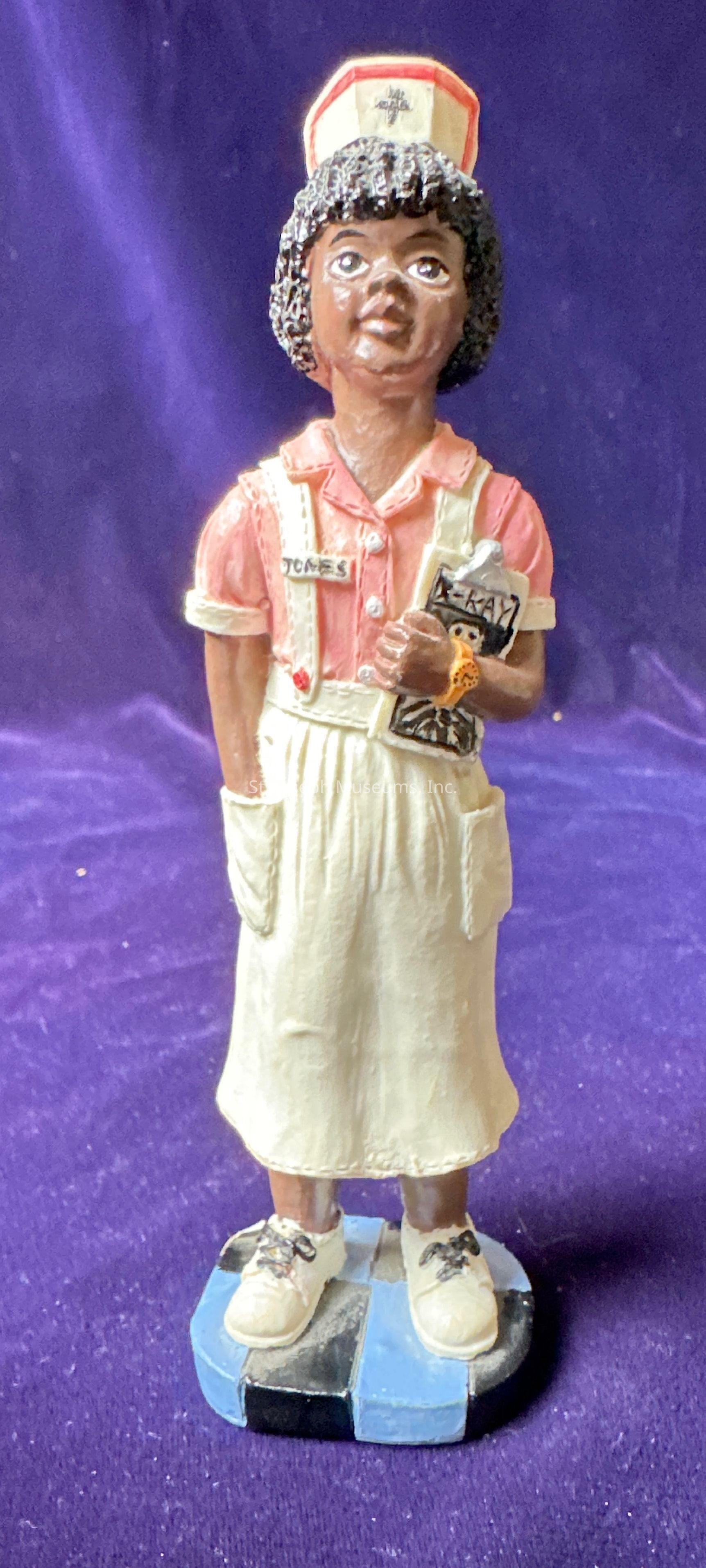 The image shows a figurine of a nurse standing on a checkered base with alternating blue and black squares. The nurse is wearing a vintage-style uniform that consists of a pink blouse with rolled-up sleeves and a white apron skirt with pockets. This nurse figurine also sports white shoes with black laces and is holding what appears to be a clipboard in one hand. She is wearing a white nurse cap with red trim, which has a medical emblem on it. The figurine is detailed, with small features such as a name tag reading "Jones" and a small watch on her wrist. The backdrop consists of a rich, deep purple fabric that adds contrast and makes the figure stand out prominently. Her facial expression is serene, and she has detailed, textured hair. The craftsmanship and attention to detail suggest this figurine is a collectible or decorative item that may serve as a tribute to the nursing profession.