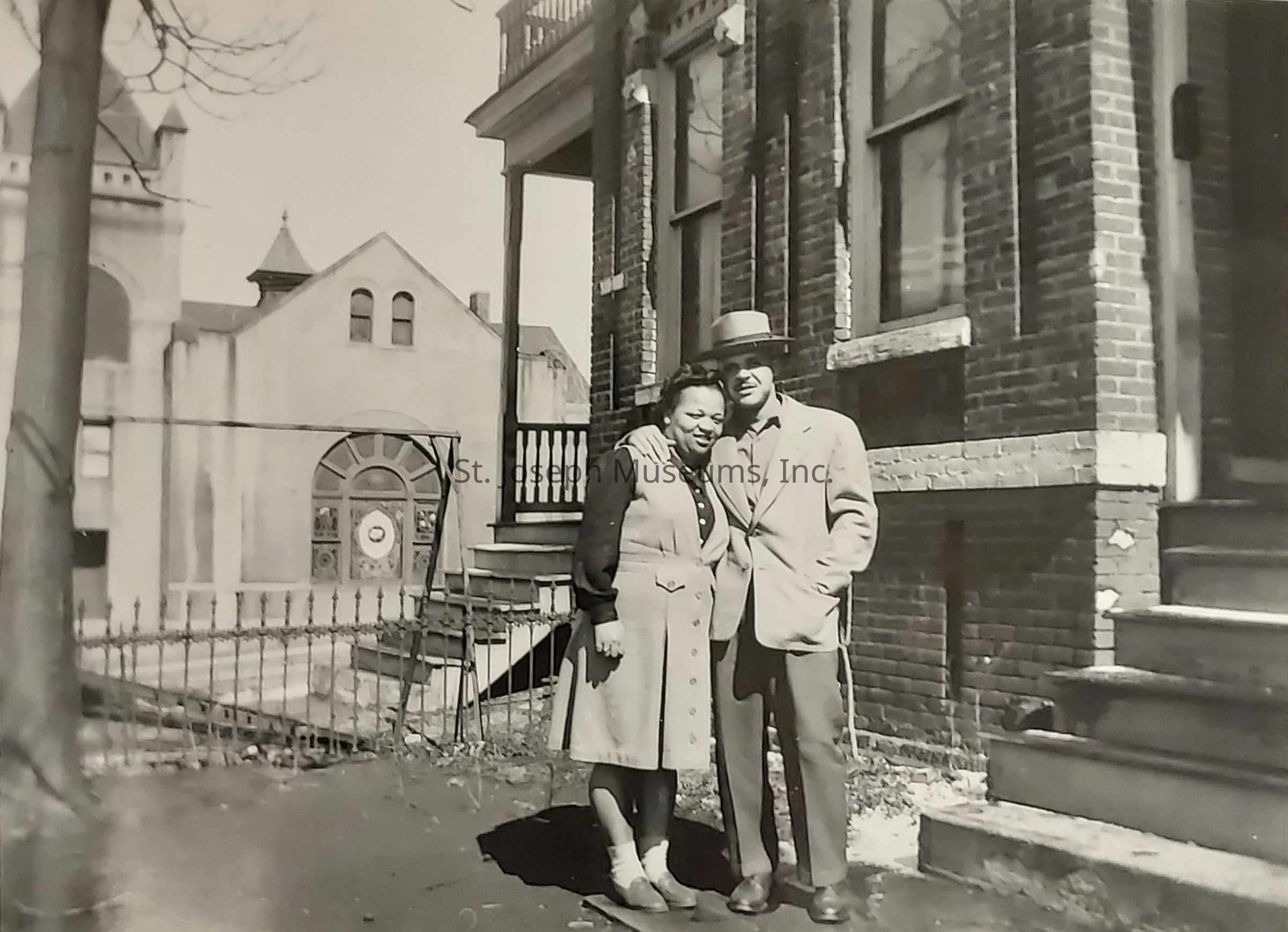 In a nostalgic black-and-white photograph, a couple stands affectionately close to each other in front of a brick building, with one person's arm gently around the other's shoulder; they are captured frozen in time during a seemingly casual moment, yet their dress suggests care and intention, as the man wears a suit and hat, while the woman wears a buttoned dress with a patterned blouse. The background reveals an urban setting with architectural details, including the iron railing and steps leading to a porch, and further back, more buildings are visible with distinctive windows and rooftops, indicating perhaps a neighborhood in a bygone era. The image conveys a sense of warmth, connection, and historic charm, capturing a slice of life from a different time period that exudes both intimacy and a broader narrative of its surroundings.