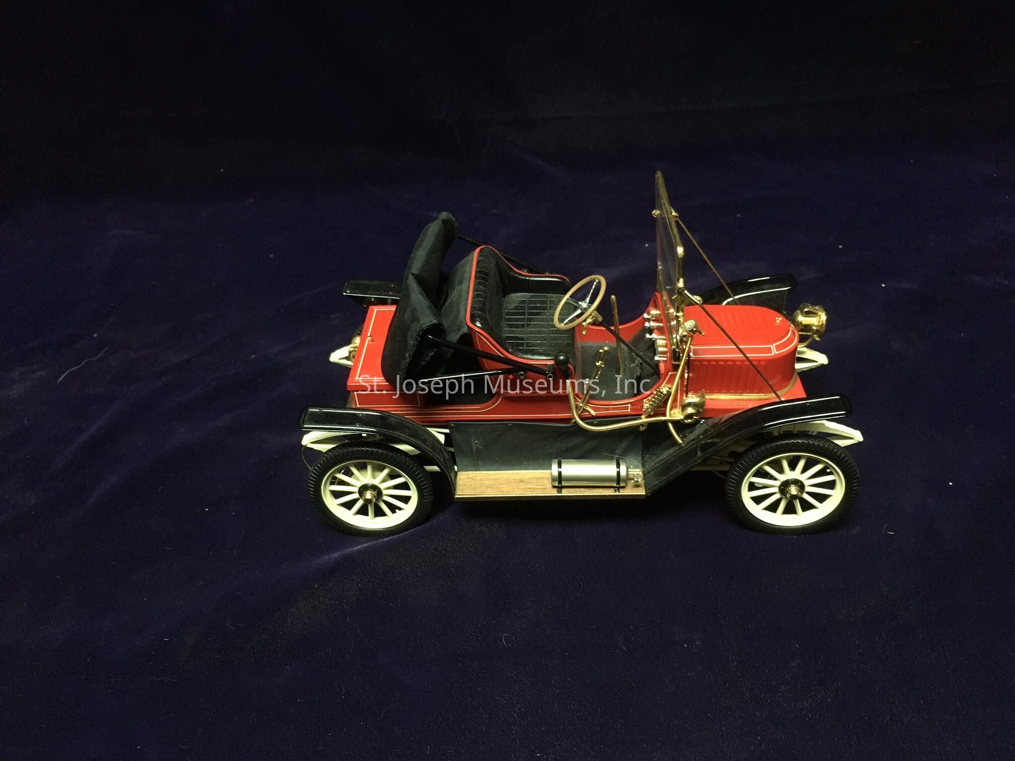The image features a finely crafted model of a vintage automobile, set against a dark background. The car showcases a striking red body with intricate black detailing, reminiscent of early 20th-century vehicles. Its open-top design includes two rows of seats upholstered in a plush black material, complemented by a stylish, thin gold steering wheel that adds an element of elegance. The wheels are white with intricate spoke designs, enhancing the overall antique aesthetic. Brass elements such as a horn and headlamp are visible at the front, further emphasizing the classic look. This model captures the essence of a bygone era, offering a glimpse into the history of automotive design. The purple-toned backdrop provides a rich contrast, allowing the vibrant colors and detailed features of the car to stand out prominently in the image.