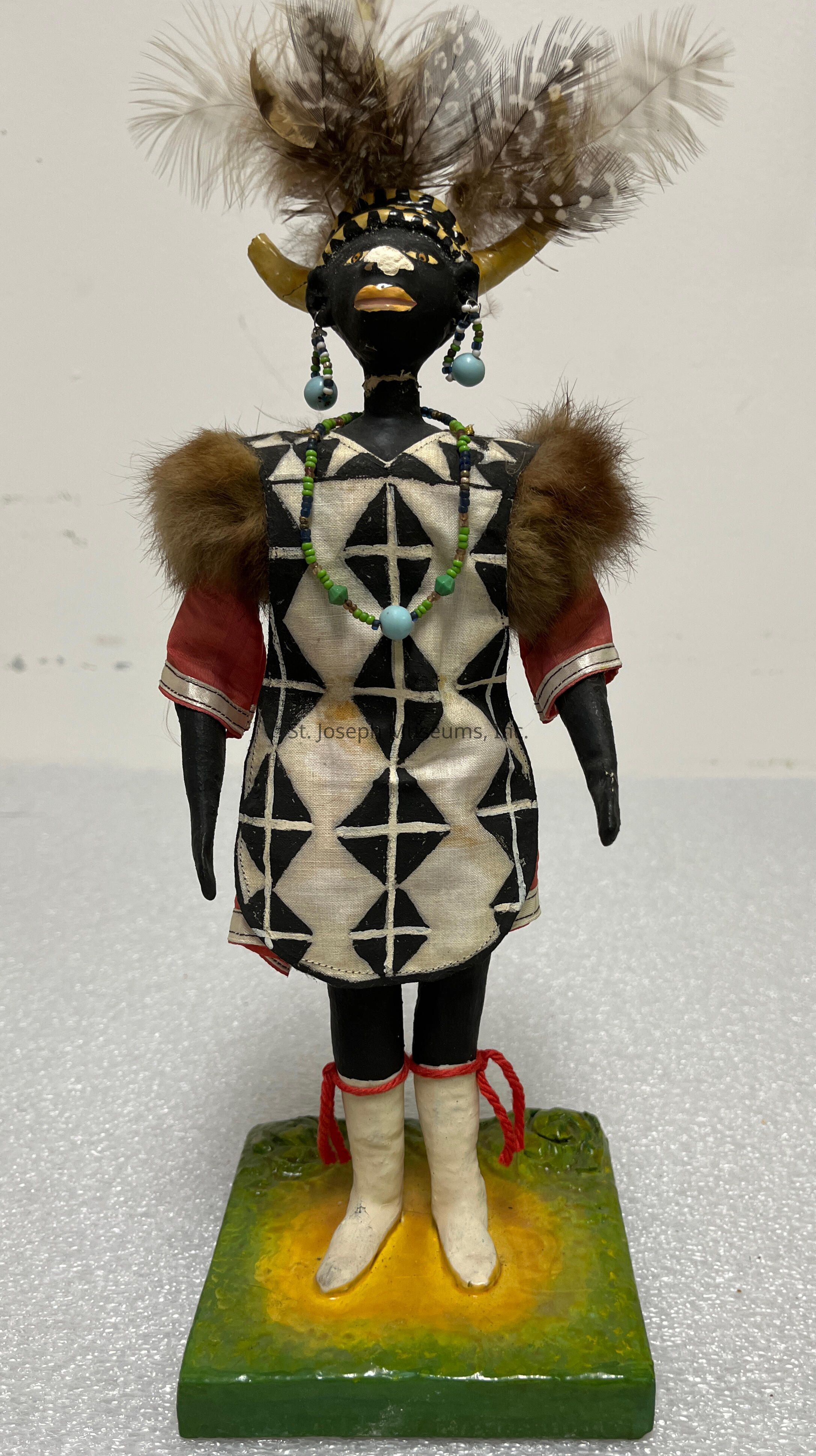The image depicts a detailed figure made from black material, dressed in a striking outfit that combines traditional and artistic elements. The figure wears a tunic featuring a bold black geometric pattern on a white background, paired with red sleeves that add contrast. Brown fur adorns the shoulders, accentuating the luxurious texture of the attire. The figure stands on a square, multi-colored base, primarily in shades of green and yellow, which adds vibrancy to the composition. Its head is adorned with an elaborate headdress featuring a collection of earthy-toned feathers and prominent curved horns, adding height and drama to its appearance. The face is painted with distinct facial markings, including a yellow-painted mouth and a white shape on the forehead, suggesting cultural significance. The figure wears decorative beaded jewelry, including a green and blue necklace and dangling blue earrings, enhancing its ornamentation. White boots with red ties complete the ensemble, contributing to the figure's unique ceremonial or artistic style.