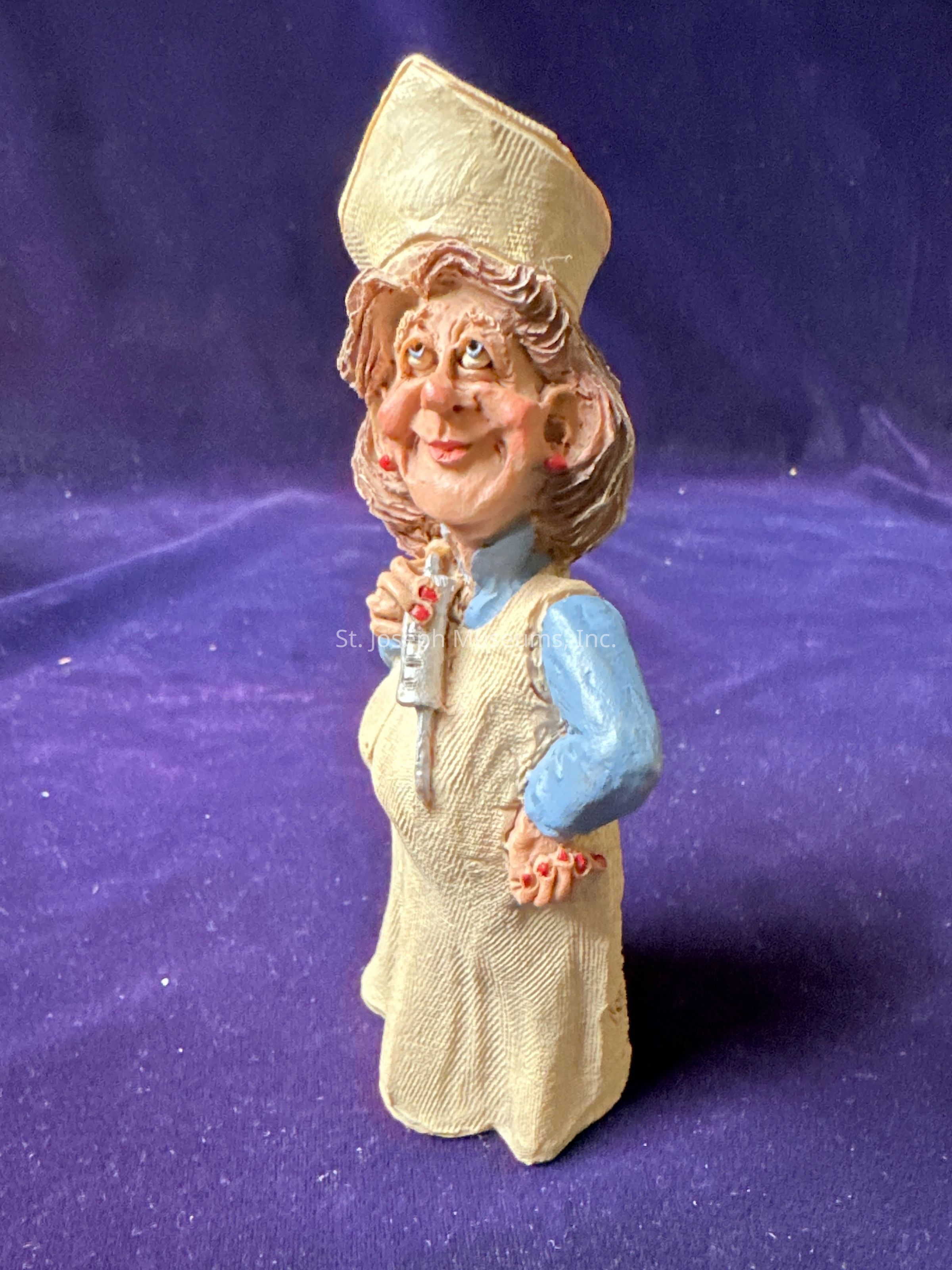 The image features a whimsical figurine of a smiling nurse set against a rich purple background. The nurse is wearing a traditional nurse's cap and a light-blue uniform with a beige apron. Her hair is styled in soft waves, and she holds a thermometer with her left hand while her right hand rests on her apron. The figurine exudes a cheerful and charming demeanor, characterized by exaggerated facial features, rosy cheeks, and a playful expression. The textured detail of the fabric in her apron adds realism, contrasting with the stylized design. The lighting creates gentle shadows that enhance the depth and dimension of this amusing and endearing piece.