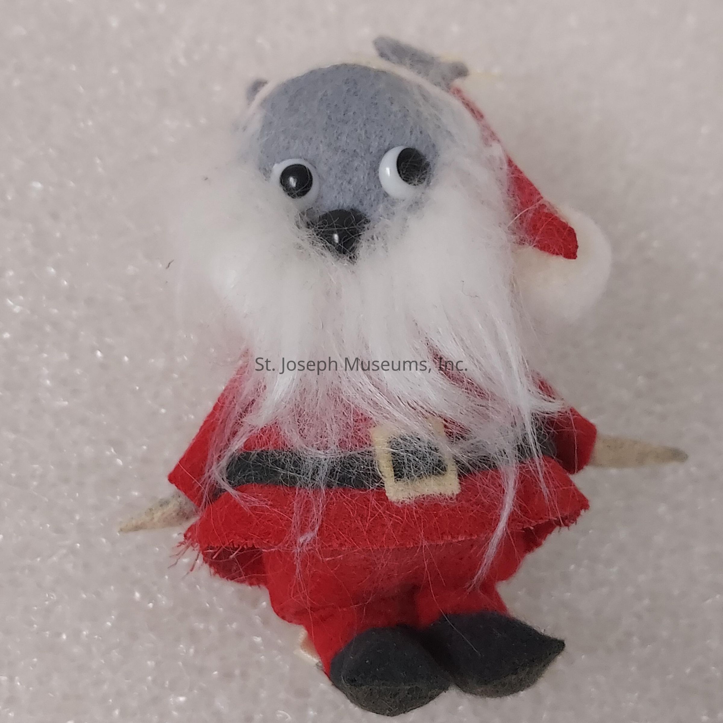 The image depicts a small, whimsical figurine resembling a Santa Claus with a unique twist; the character has a grey dog-like face with a black nose and is adorned with googly eyes, giving it an amusing expression; it features a fluffy white beard and dons the traditional red-and-white Santa outfit, complete with a black belt with a yellow buckle and matching boots, while resting on a textured, light background, possibly resembling snow, which adds to the festive, playful theme of the figure, likely serving as a charming decoration piece for the holiday season, and showcasing a blend of creativity and humor.