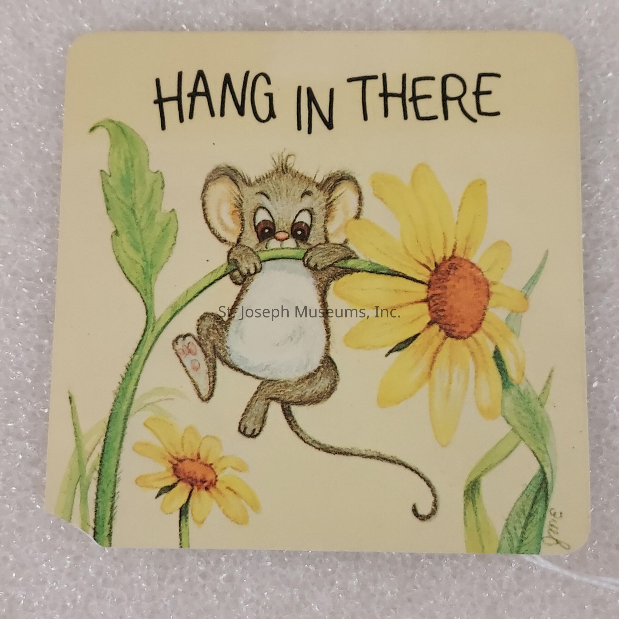 The image depicts an illustrated square card with a soft pastel background, featuring an adorable cartoon mouse clinging to the stem of a large, bright yellow flower with a brown center, surrounded by green leaves and another smaller flower beneath it. The mouse, with large ears and an expressive face, appears playful and cheerful, its little feet gripping the plant as its long tail curls below, symbolizing resilience and tenacity. Above the scene, the bold, block-lettered text “HANG IN THERE” conveys a motivational message, exhorting viewers to persevere through challenges.