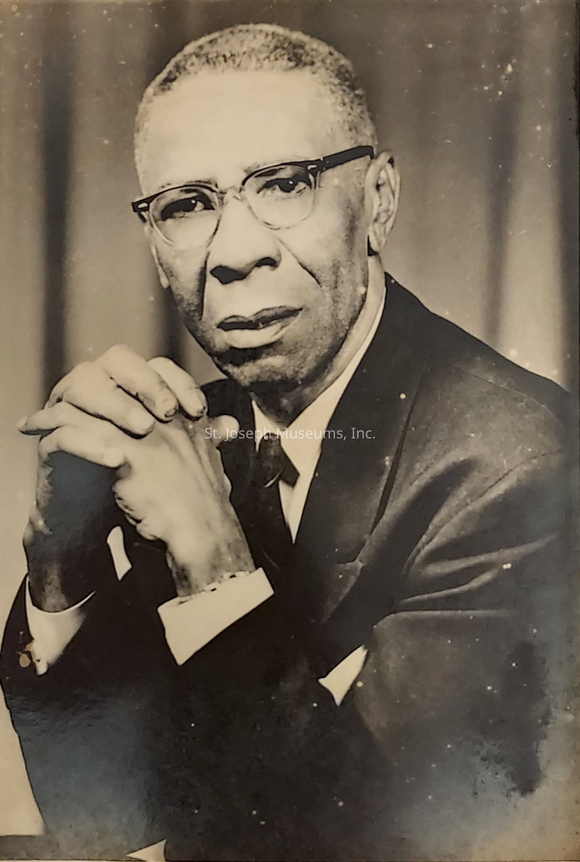 The image is a black and white photograph depicting an older gentleman dressed in a formal suit and tie, wearing glasses, and gazing intently at the camera with a serious, thoughtful expression; his hands are clasped together beneath his chin, conveying a sense of contemplation or introspection; the background is blurred, directing focus toward his face, and there is a classic, timeless quality to the photograph, suggesting it may have been taken several decades ago, which adds to its historical or vintage feel.