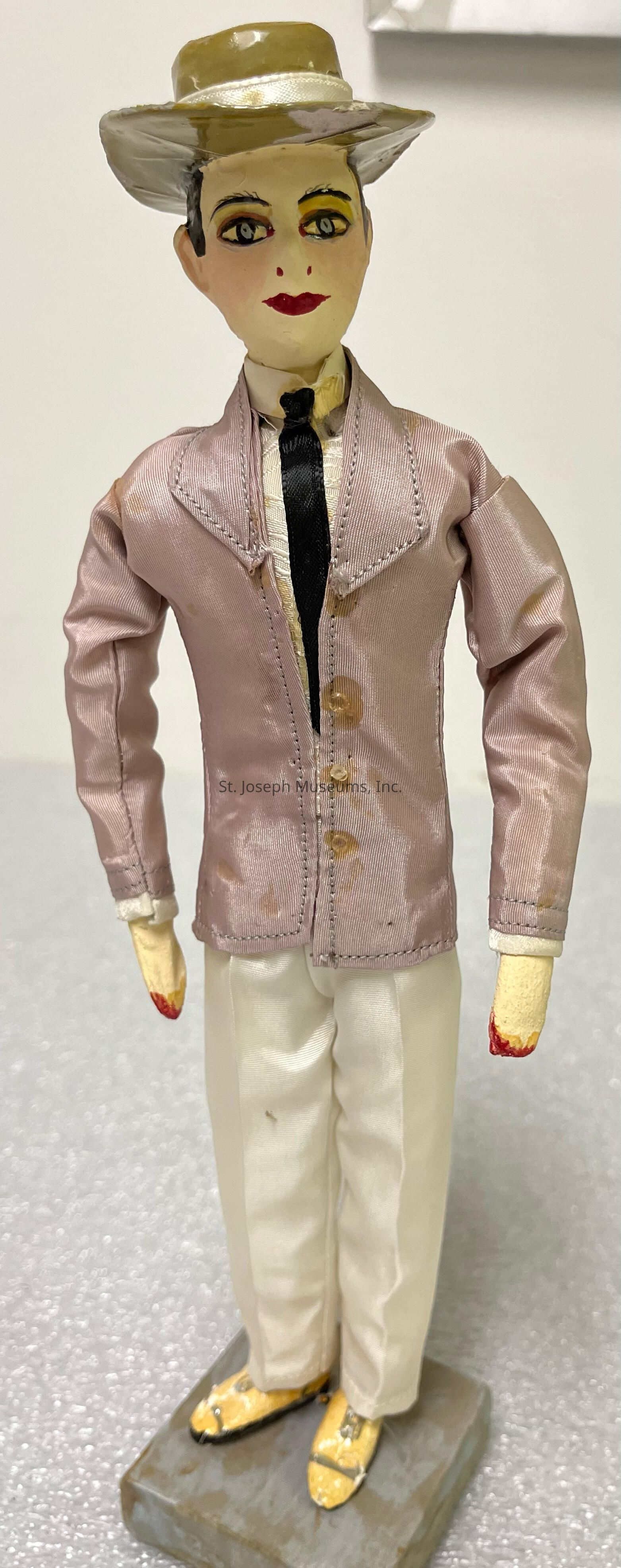 The image features a handcrafted doll dressed in vintage attire, showcasing an elegant and retro look reminiscent of early 20th-century fashion. The doll is adorned with a wide-brimmed hat, embodying a stylish flair with its woven texture and ribbon accent. Its facial features are delicately painted, with expressive eyes and bold red lips bringing a lifelike quality to its appearance. The doll's outfit consists of a finely tailored jacket in a soft pink hue, fastened with small buttons, over a crisp white inner shirt and a black tie, all emphasizing a formal yet fashionable theme. Completing the ensemble are white trousers that extend down to the doll's painted shoes, placed on a sturdy base, which helps the figure stand upright. This artistic creation, with its attention to detail and nostalgic elegance, captures the essence of a bygone era, mirroring the aesthetics and sophistication of that period while serving as an intriguing collector's item or a charming decorative piece.