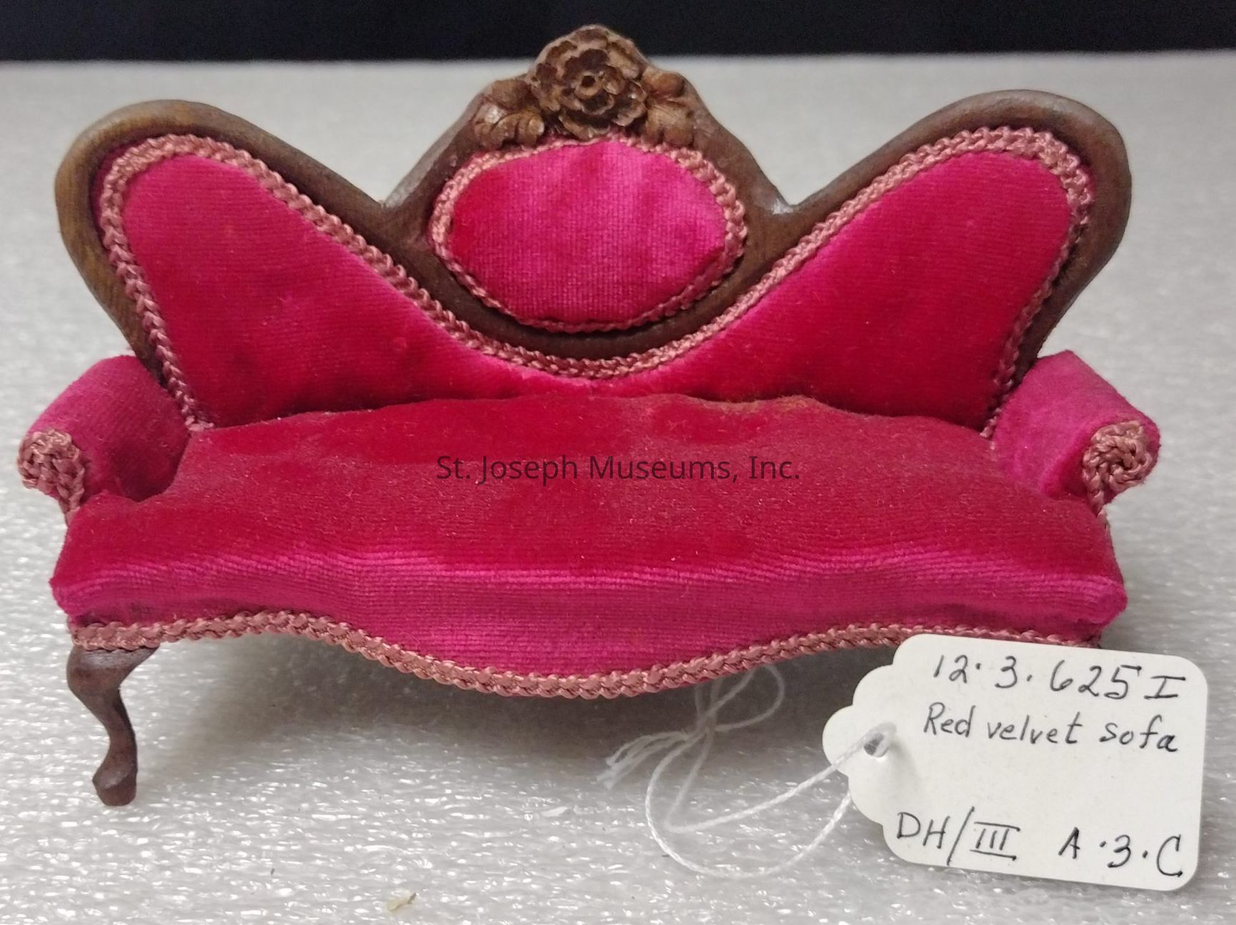 The image depicts a small, ornate sofa upholstered in rich red velvet, featuring an intricately carved wooden frame with a floral design at the top of its high backrest. The sofa exhibits classic Victorian styling with its curved arms and elegantly arched back, providing an impression of elegance and refinement. It is positioned on a textured surface, and a white tag is attached to one of its legs. The tag contains handwritten text, reading "12.3.625I Red velvet sofa DH/III A.3.C," suggesting it may be part of a collection or an exhibit. The luxurious velvet fabric gives the sofa a plush appearance, enhanced by the braided trim that frames the seating area and backrest. This piece combines artistry with craftsmanship, indicative of a time when intricate detailing was prevalent in furniture design. The overall effect is one of opulence and sophistication, capturing the viewer's attention with its vibrant color and detailed ornamentation, standing as a beautiful representation of vintage design and style.