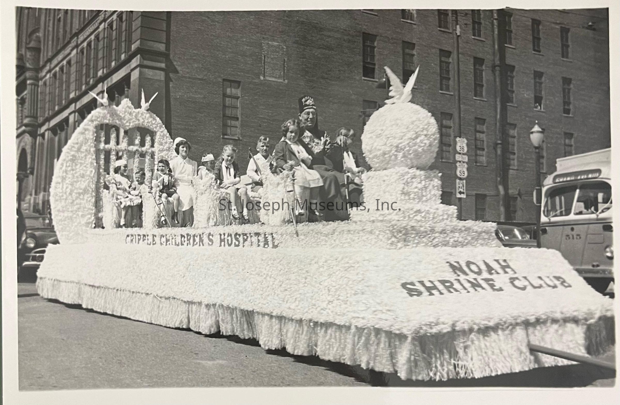 Black-and-white photograph depicting the Shrine Crippled Children's Hospital float during an Apple Blossom Parade in St. Joseph, Missouri. The float, adorned with intricate decorations and featuring children and adults seated, rolls through a street lined with brick buildings. Signs on the float read "Cripple Children's Hospital" and "Noah Shrine Club."
