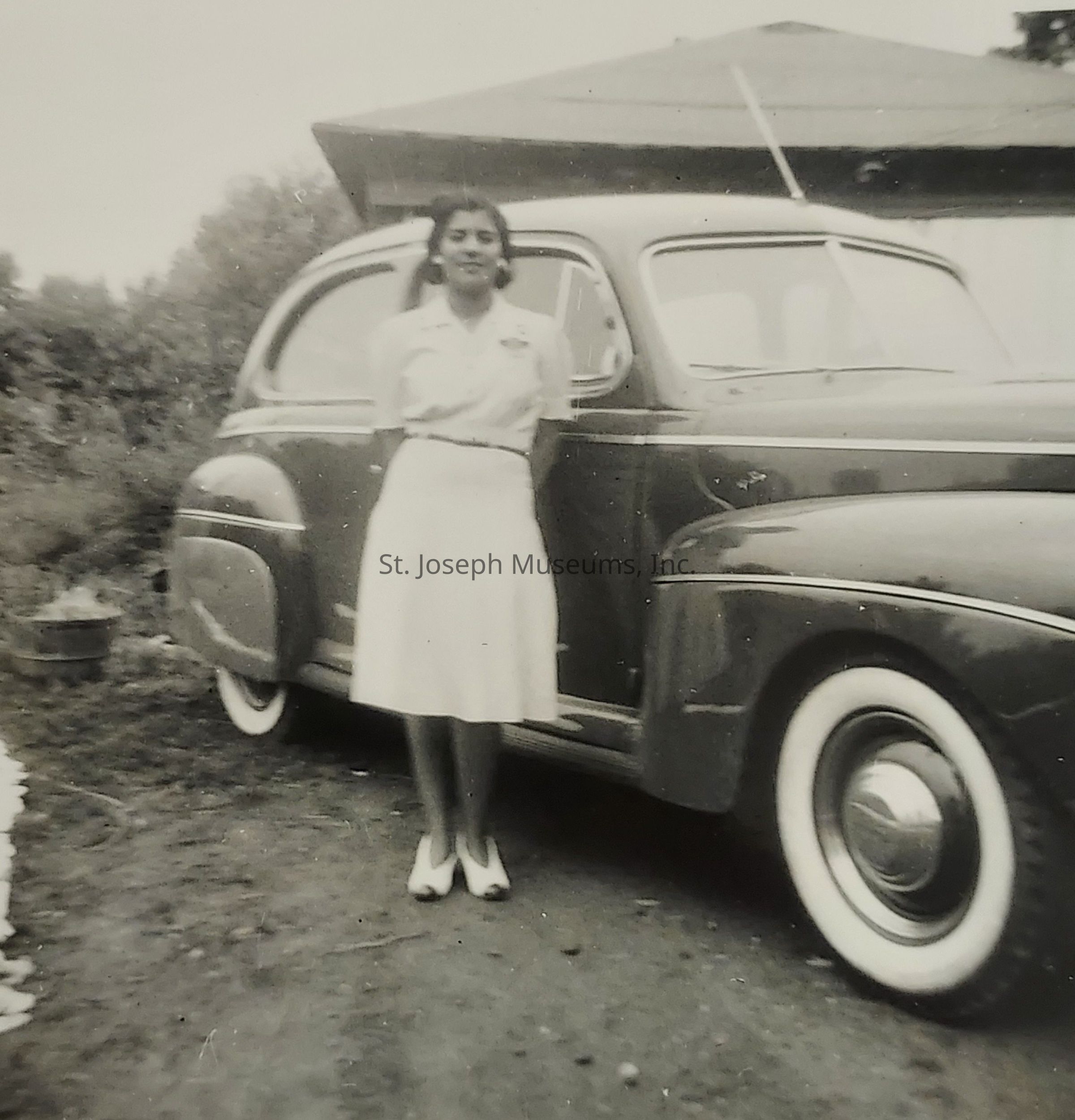 In this black-and-white photograph, a woman is standing in front of a vintage car, possibly from the mid-20th century, wearing a light-colored blouse and a skirt, complemented by white shoes, with her hands behind her back. The car, with its classic rounded design and prominent white-wall tires, is parked on a patch of dirt near a modest house. To the side, a small planter pot can be seen, hinting at a garden setting, while the background shows trees that suggest a rural or suburban environment, reflecting a nostalgic slice of life from a bygone era.