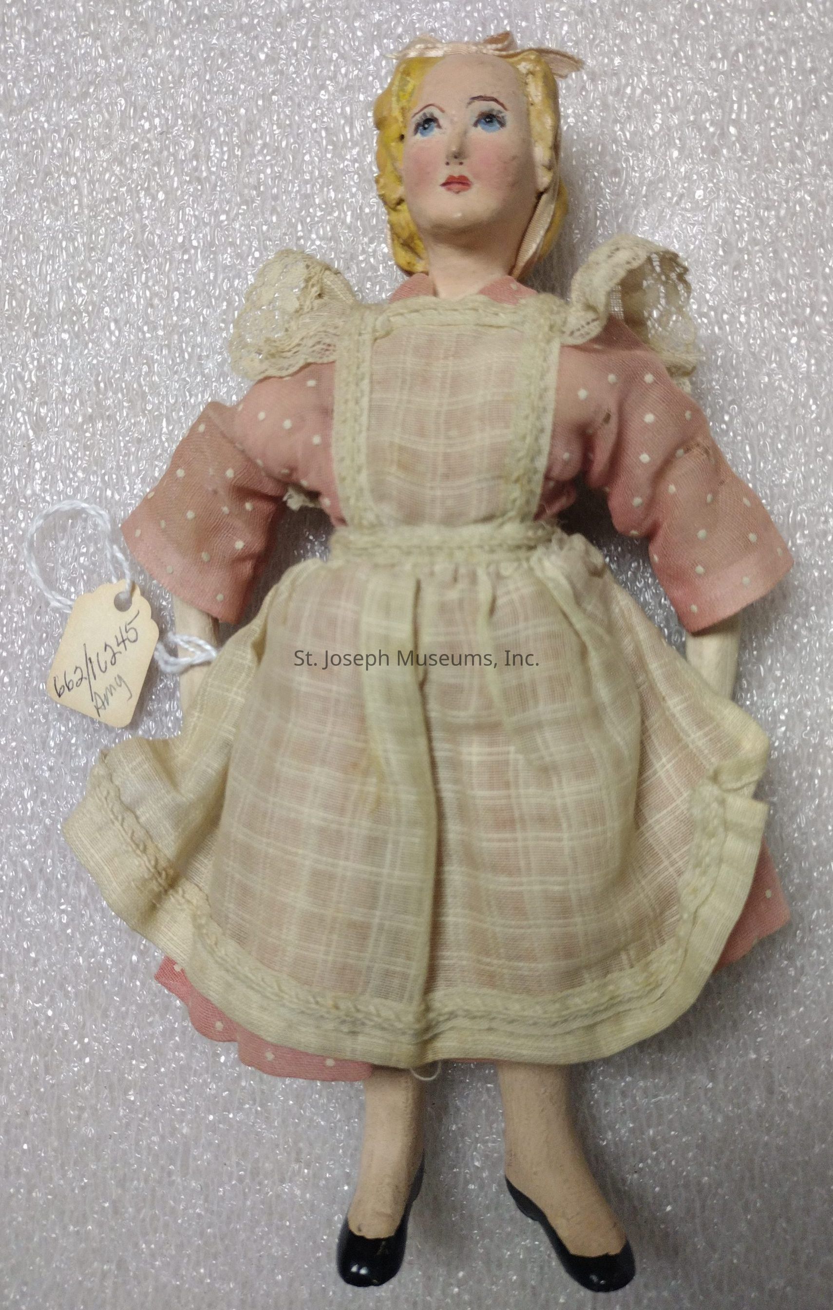 The image shows a vintage-style doll with a porcelain-like face featuring delicate, painted features, including light blue eyes, rosy cheeks, and red lips. It has blond, curly hair styled neatly, and wears a pale pink dress adorned with white polka dots and lace detailing around the collar. Over the dress, the doll sports a cream-colored, checkered apron, which is tied at the back. Its arms are slightly bent, with visible stitching. The doll's legs are covered with stockings and it wears small black shoes. A tag with numbers and the word “Amy” is attached to one arm, suggesting it may be a collector's item or part of an exhibition. The background is a textured white surface that accentuates the doll’s vintage aesthetic, creating an antiquated and charming appearance.