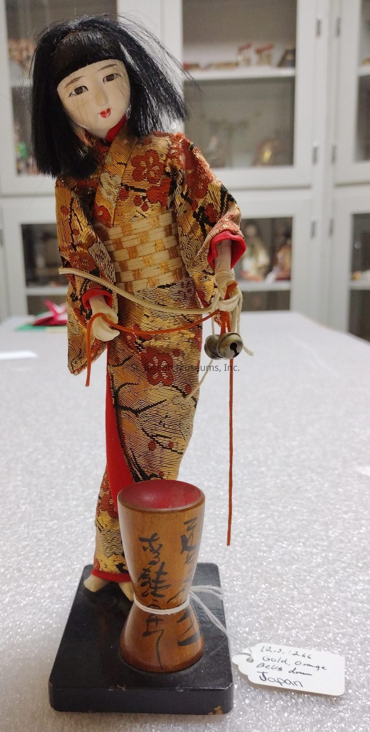 The image depicts a traditional Japanese doll featuring a delicate face and black hair styled in a bob. The doll is dressed in an elaborate gold and red kimono adorned with intricate floral patterns, reflecting the elegance of traditional Japanese attire. In its hand, the doll holds a string attached to a small bell, adding an element of charm. The doll stands on a wooden base, which has Japanese characters inscribed on it, contributing to its cultural authenticity. A tag attached to the base notes its origin as Japan, and mentions "gold, orange, bell, drum," possibly describing the materials or themes of the doll’s attire and accessories. The background consists of a softly focused set of cabinets, suggesting the setting might be a display or a store showcasing cultural or artistic items. The foreground, where the doll is placed, appears to be covered in a white textured fabric, providing a simple yet effective contrast that emphasizes the detailed design of the doll.