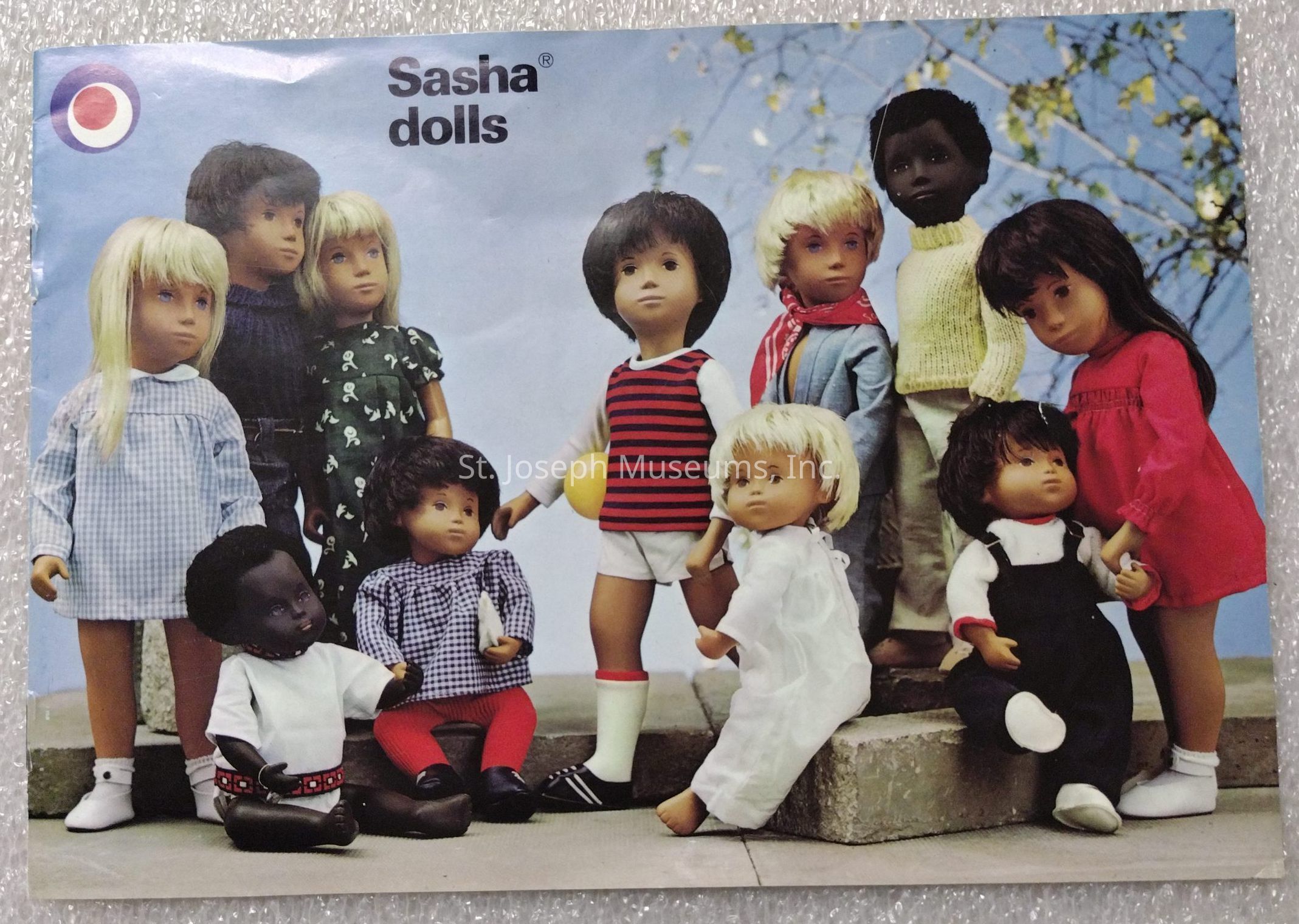 The image features a group of Sasha dolls, each with distinct clothing, hairstyles, and skin tones, arranged closely together against a soft background with a hint of tree branches. The dolls display a variety of outfits, including checkered dresses, striped shirts, knit sweaters, and solid-colored garments. Some dolls are standing while others are seated on a concrete step, suggesting a casual, friendly gathering or playtime scene. The diversity in the dolls' appearances reflects an intention to represent different ethnicities and styles, characteristic of Sasha dolls' inclusive design. The overall atmosphere is cheerful and inviting, with the dolls exuding a sense of camaraderie and playfulness, enhanced by the clear, bright lighting that emphasizes their expressions and the colorful details of their attire. The cover's title, "Sasha dolls," is displayed prominently at the top, setting the thematic focus of the image.