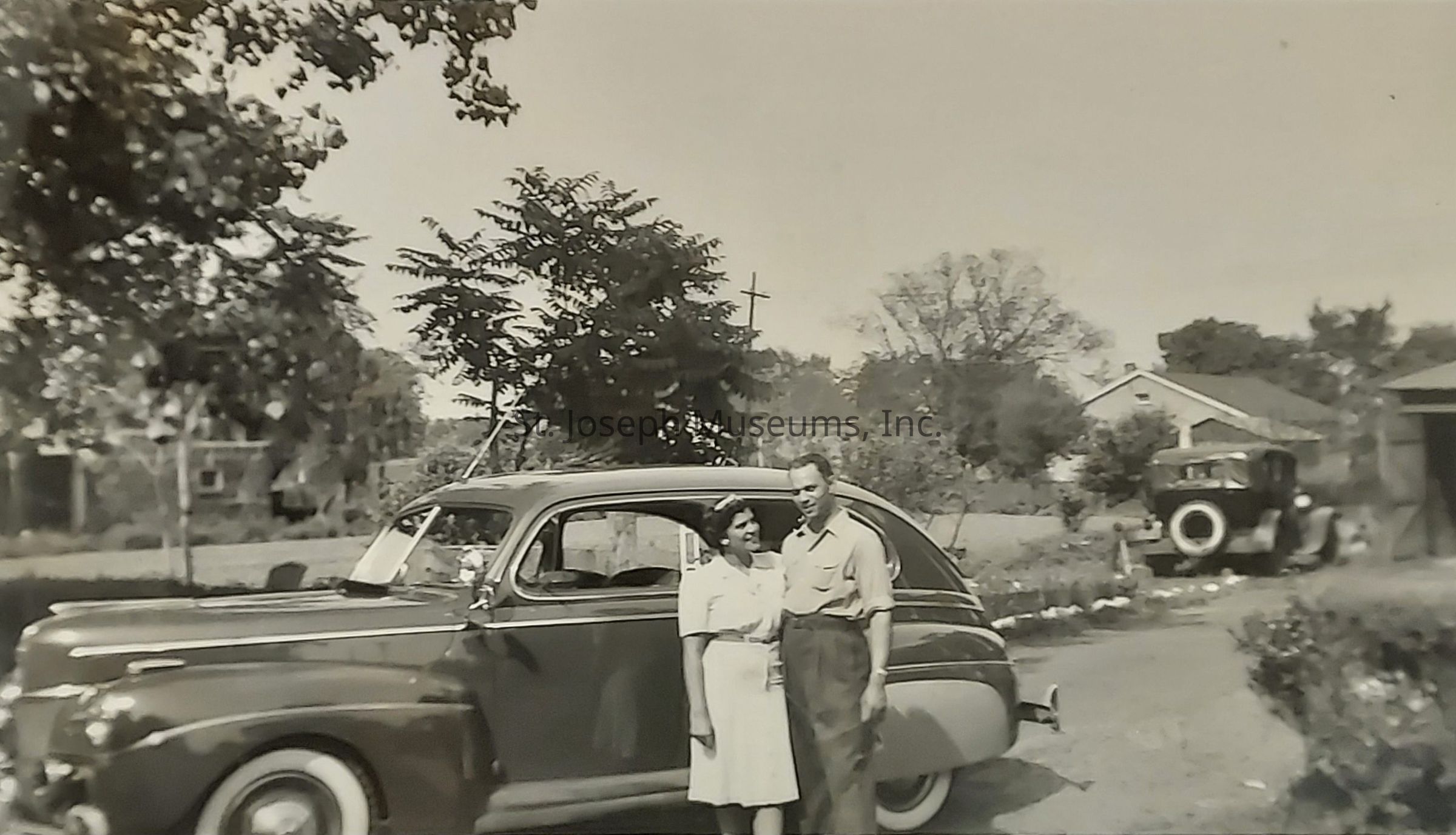 The black and white image captures a nostalgic scene from the past, showcasing a couple standing affectionately beside a classic car, likely from the mid-20th century, amidst a serene suburban setting. Tall trees frame the background, their branches casting delicate, dappled patterns of light and shadow on the ground. A quaint house with a pitched roof is visible in the distance, hinting at a peaceful, family-friendly neighborhood. In the far background to the right, another vintage car is parked, echoing the era’s automotive design trends with its prominent rounded body and whitewall tires.