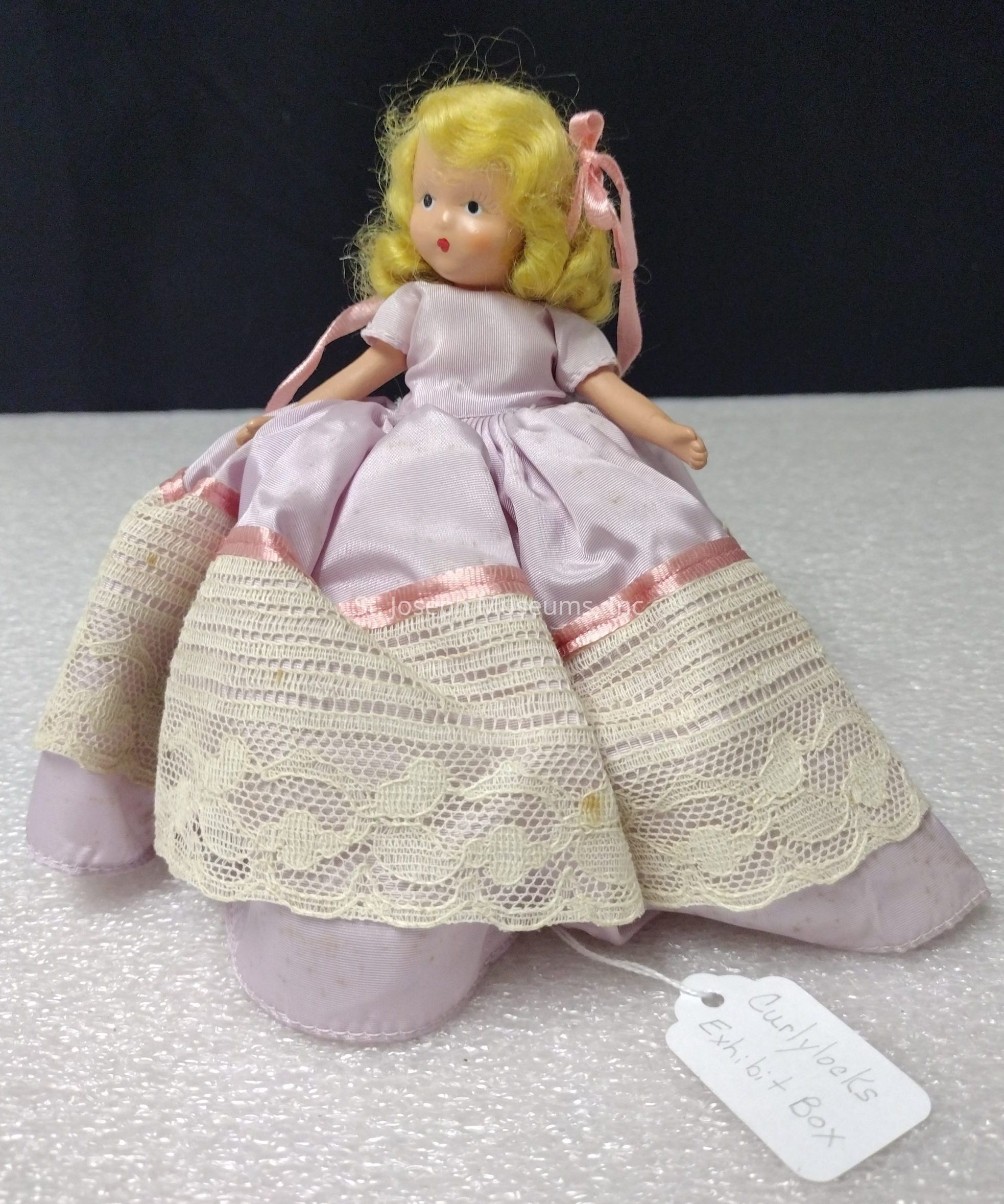 The image shows a vintage doll with blond curly hair adorned with a pink ribbon, wearing a pastel pink dress featuring delicate lace and satin trim. The lace detail on the skirt is intricate, emphasizing a sense of craftsmanship. The doll is seated on a textured white surface against a dark backdrop, bringing attention to the soft colors and details of its attire. Dangling from the doll's dress is a small tag that reads "Curlylocks Exhibit Box," suggesting it may be part of a collection or display. The overall composition creates an impression of nostalgia and charm.