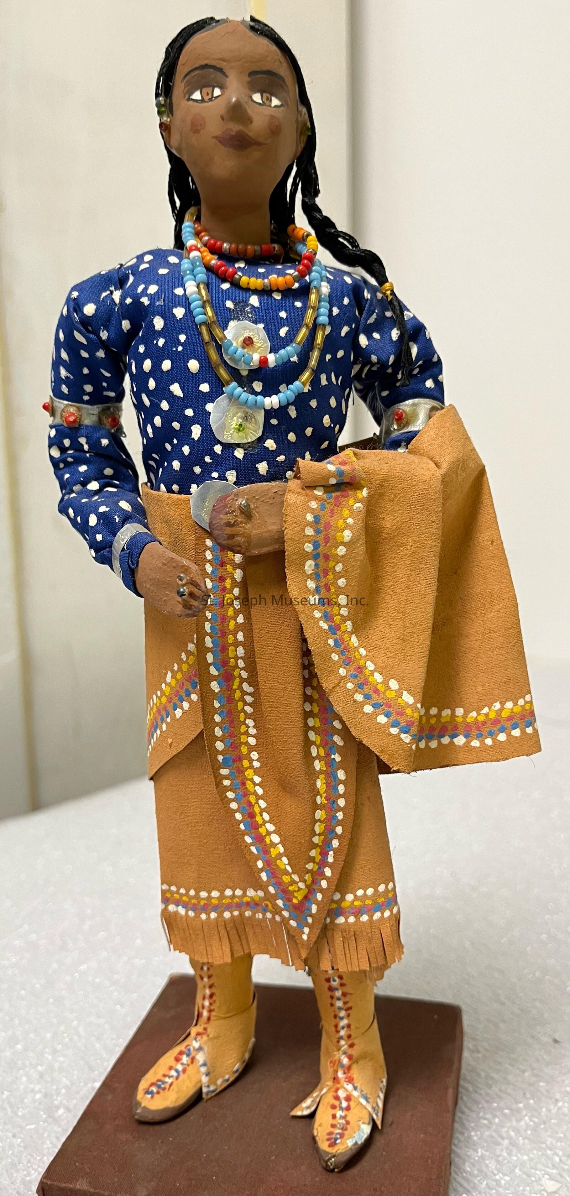 The image depicts a handcrafted doll dressed in traditional attire; it features a detailed blue blouse with white polka dots, layered multicolored beaded necklaces, and an ornate brown skirt with painted patterns in blue, red, yellow, and white. The figure has black braided hair. Its brown fringed boots complement the overall outfit, while an additional fabric piece is draped over one arm.