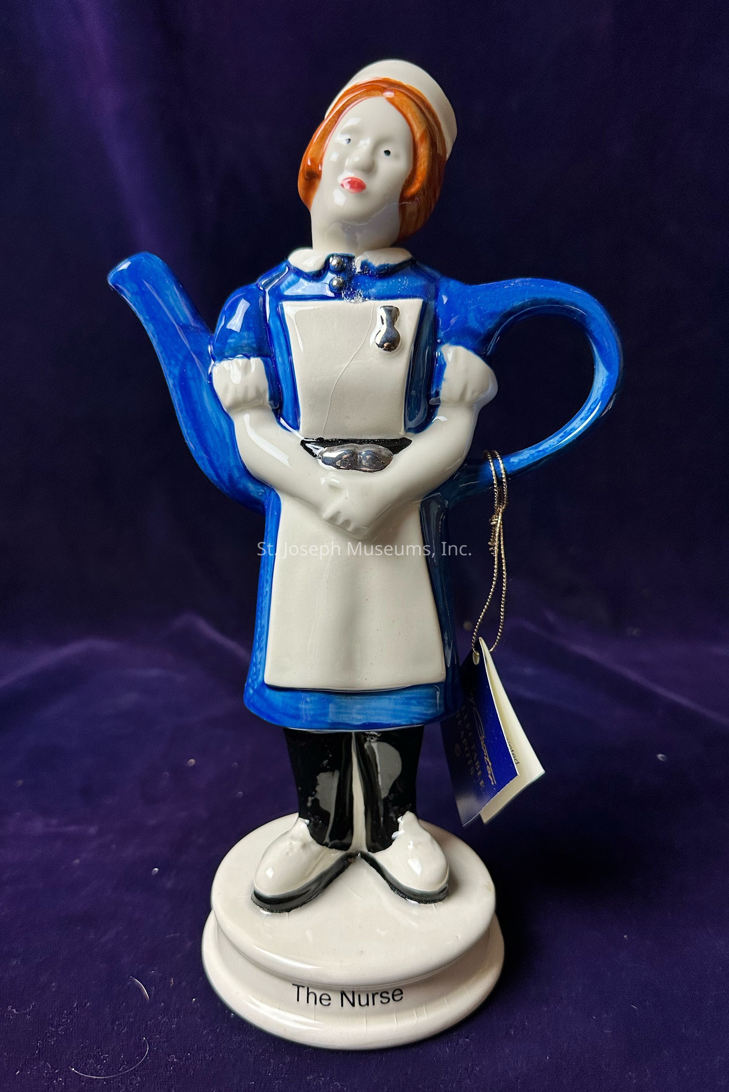The image depicts a creatively designed teapot shaped like a nurse, featuring a blue uniform with white apron detailing, complete with a cap. The spout of the teapot resembles an arm, forming the overall figure's profile, while the handle mimics the other arm, positioned on the hip. The teapot stands on a circular base labeled "The Nurse" in black lettering. The nurse figure has red hair and is painted with a calm expression, creating a whimsical and artistic representation. It stands against a rich, velvet-like purple background, enhancing the vibrancy and detail of the teapot's colors.