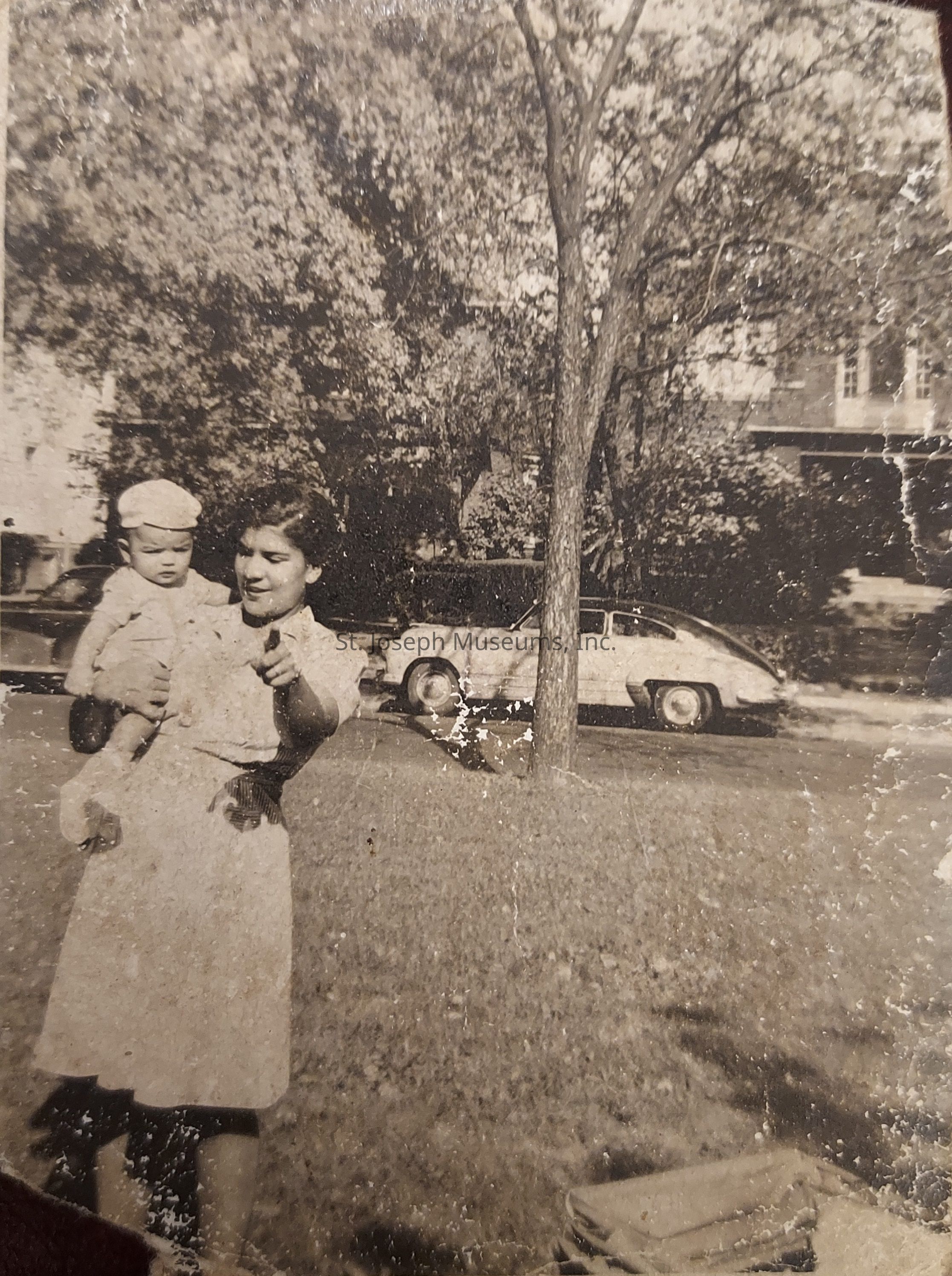 In this vintage black and white photograph, a woman is holding a child while standing on a grassy area, with tall trees and a parked classic car in the background, capturing a moment in time that suggests a setting from mid-20th century suburban life. The woman is smiling and pointing at something off-camera, adding a sense of interaction and liveliness to the image. The child, who is wearing a cap and a matching outfit, looks away with curiosity. The presence of the old-fashioned car and the style of clothing provide a nostalgic view into the past, evoking a sense of history and familial bonds. The trees are full of leaves, indicating that it might be spring or summer. The picture shows signs of wear, with scratches and creases, underscoring its age and the countless memories it holds. Overall, the photograph exudes warmth and simplicity, highlighting a candid moment shared between the woman and the child, layered with the charm and character typical of bygone eras.