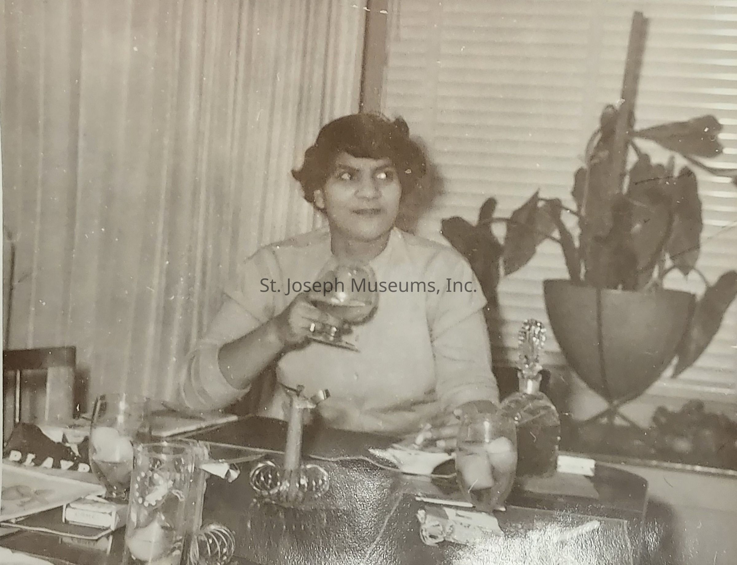 The black-and-white photograph captures a woman seated at a table, holding a large glass, possibly containing a beverage, as she gazes off to the side with a thoughtful expression, set against the backdrop of a room adorned with a large potted plant and horizontal blinds, creating a cozy and intimate atmosphere; the table in front of her is neatly arranged with various items, such as a small ornamental stand, a glass filled with ice, and a bottle, suggesting a scene of leisurely dining or social gathering, where the presence of a soft curtain on one side and the arrangement of objects lend an air of sophistication to the setting, evoking a sense of nostalgia and mid-20th-century ambiance, highlighted by the woman’s classic attire, her hair styled in soft waves, and subtle details like rings on her fingers, all contributing to a timeless portrayal of elegance and quiet contemplation.