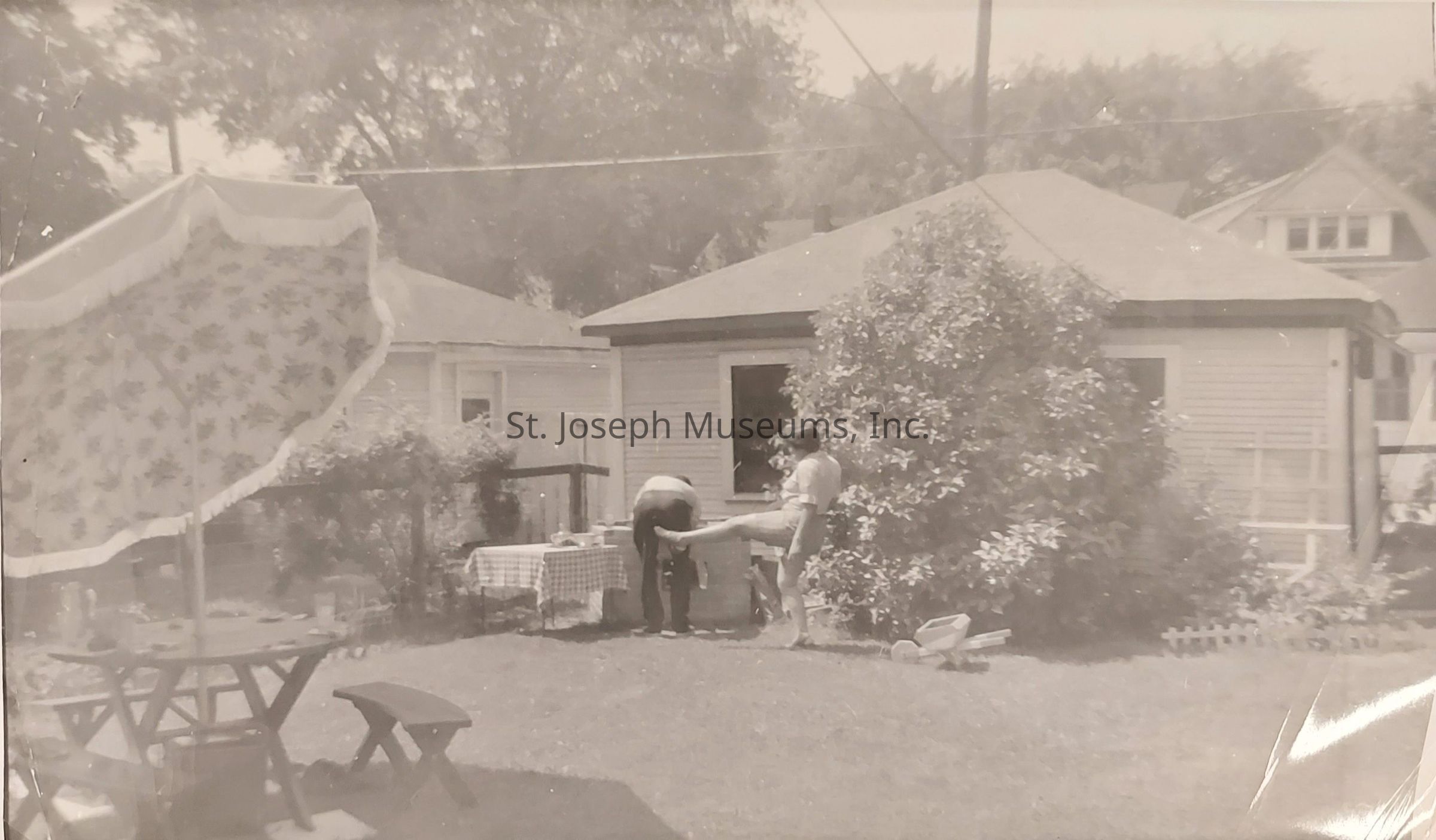 In the black-and-white photograph, a backyard scene unfolds on a sunny day, featuring two individuals near a small building adorned with horizontal siding and a visible window, surrounded by lush greenery including a large bush. One person leans forward over a table covered with a checkered cloth, seemingly engaged in an outdoor activity, while the other stands nearby with one leg bent, creating a sense of casual interaction. To the left, a large umbrella with a floral pattern provides shade over a wooden picnic table and benches, evoking a relaxed, quaint atmosphere. The yard includes a well-maintained lawn, and houses with peak roofs are visible in the background, suggesting a suburban setting from an earlier era.