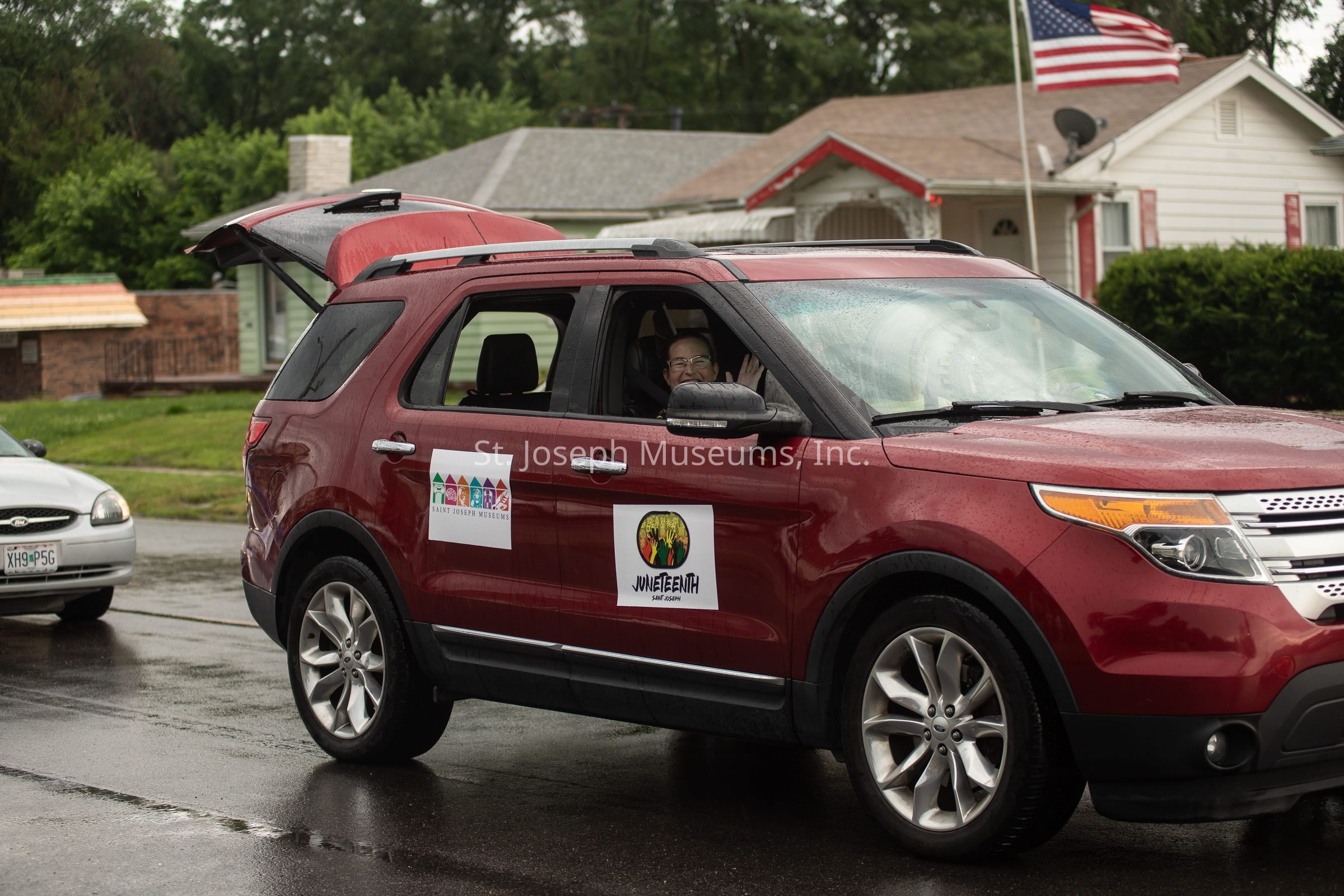 In the image, a red SUV is parked on a wet street with its rear hatch open, and features two distinct signs on its side; one promoting "Juneteenth" and the other labeled "St. Joseph Museums." A person, visible through the open passenger window, is smiling and waving towards the camera. The surroundings include another parked vehicle, a grassy lawn, and a few residential buildings in the background. An American flag can be seen on a pole beside one of the houses, lending a sense of community to the scene. The overcast weather is evident from the shiny, rain-soaked surface of the road.
