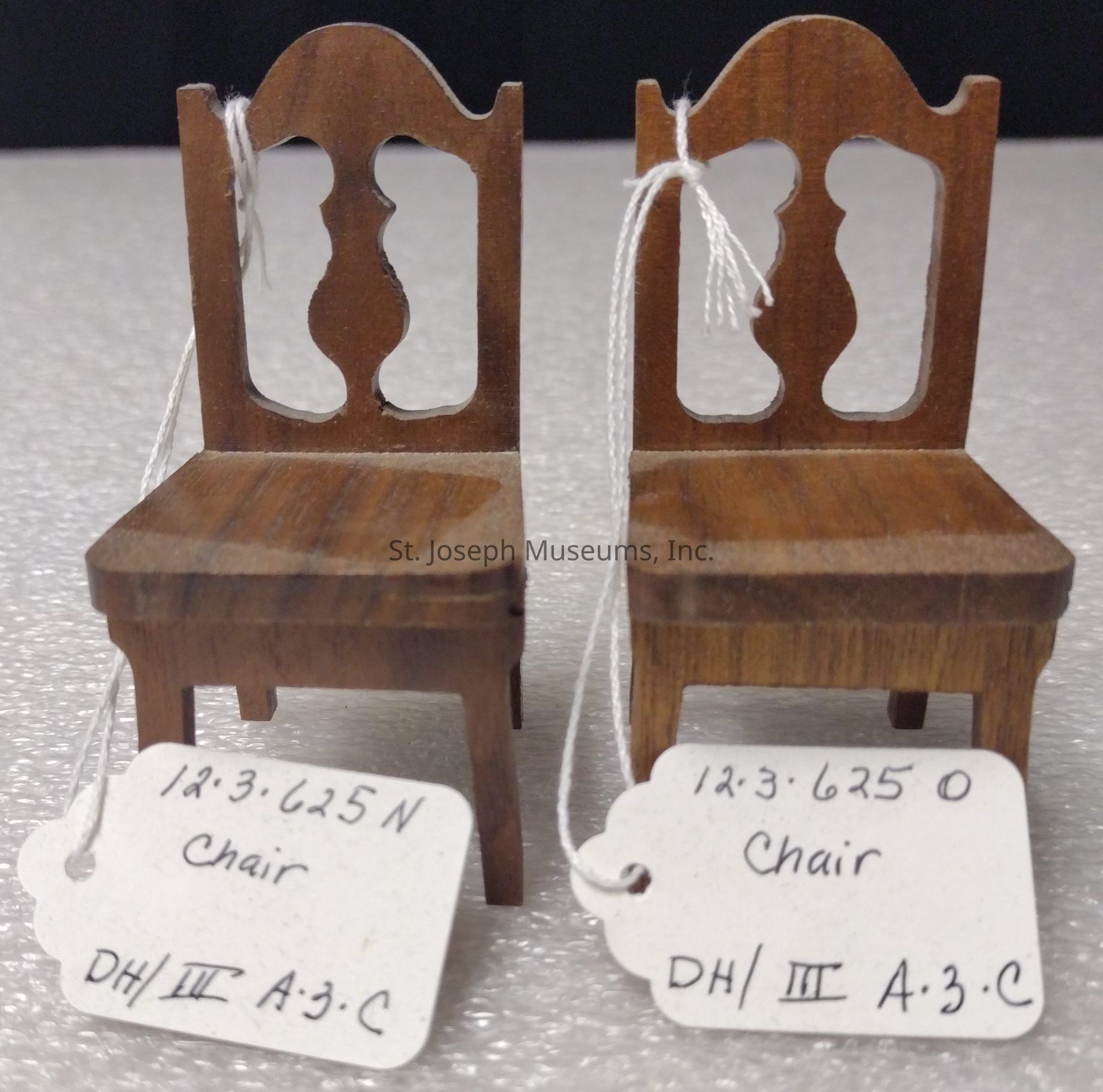 The image depicts two small, intricately crafted wooden chairs placed side by side on a textured surface. Both chairs feature an elegant cut-out design in the backrest, resembling traditional wooden craftsmanship. Attached to each chair is a white tag, with handwriting detailing information such as "Chair" and seemingly catalog numbers. These numbers read "12.3.625N" and "12.3.625O," and there are additional markings including "DH/III A:3:C." The chairs have a glossy finish, highlighting the rich wooden texture. The tags are tied with a white string around the backrest, indicating that these chairs might be part of a collection or display.