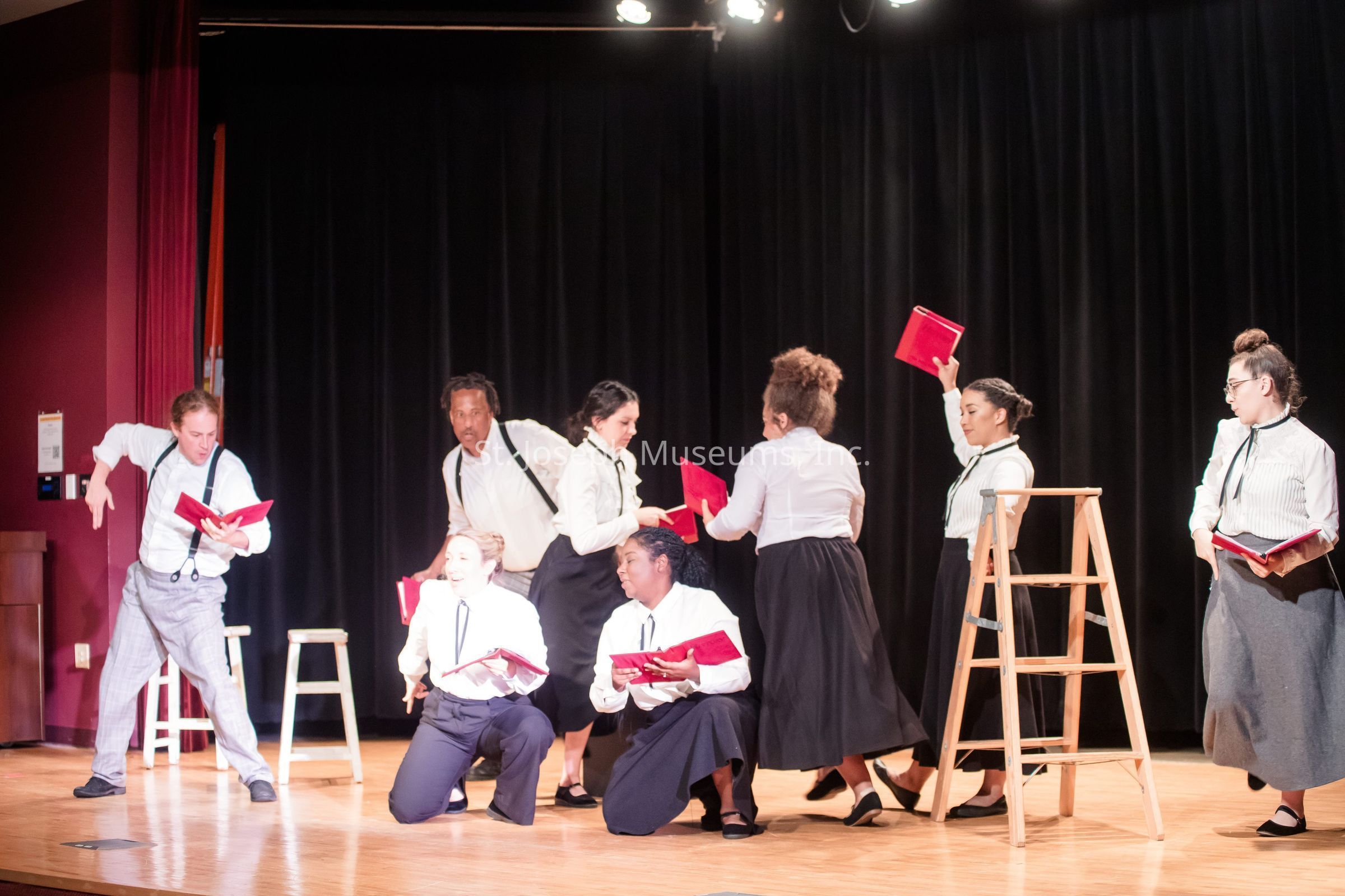 The image depicts a theatrical scene on a stage where a group of eight performers, dressed in coordinated attire consisting of white shirts and dark trousers or skirts, engage in an energetic performance. They hold red books which appear to be part of the act, suggesting a dynamic reading or storytelling session. The performers exhibit expressive body language, some standing, some crouched, creating a vivid sense of motion and interaction among them. A wooden ladder is present onstage to the right, adding an element of the set. The stage is lit brightly, contrasting with the dark backdrop, drawing all attention to the performers' animated expressions and lively gestures.