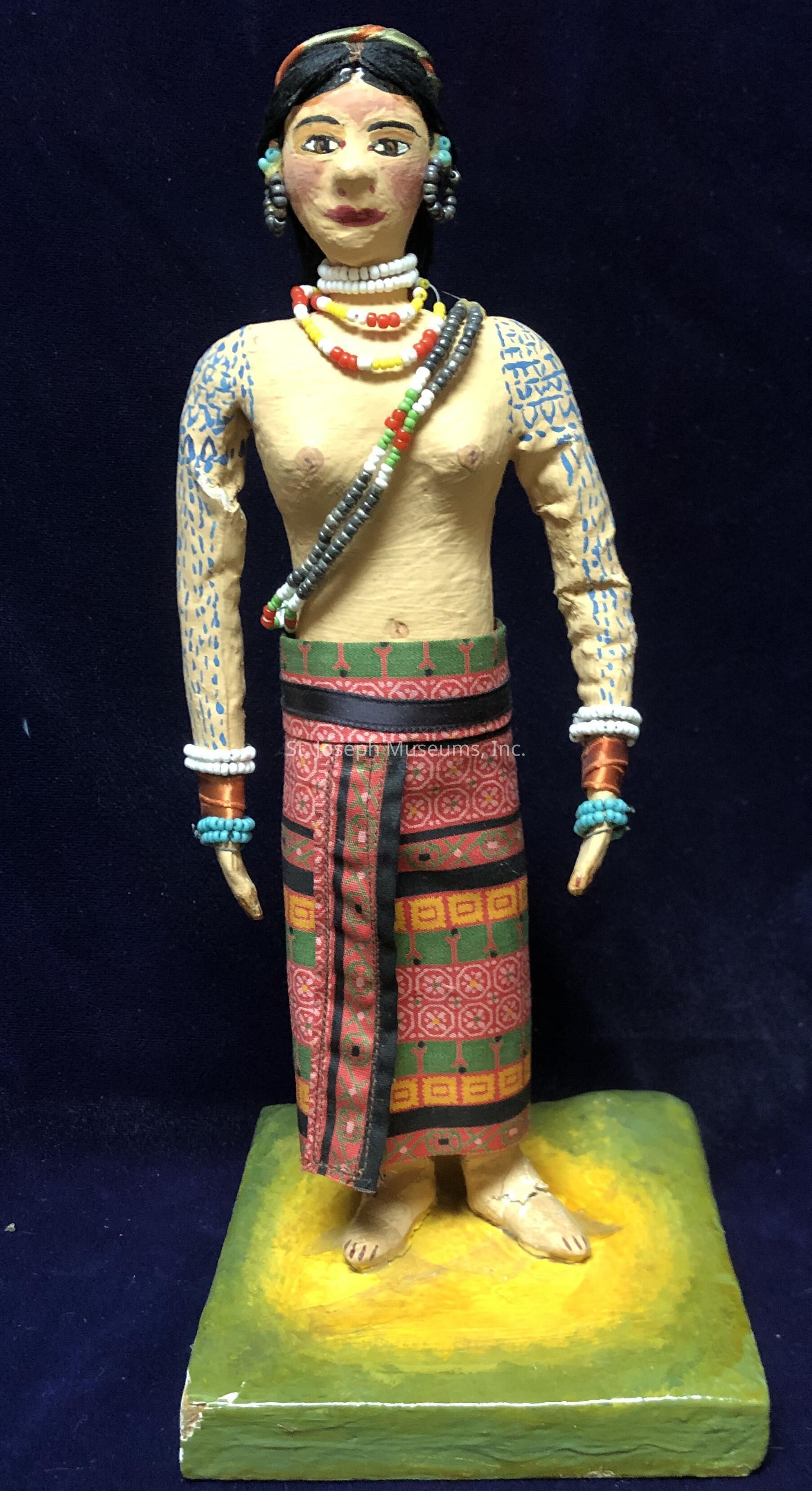 The image depicts a handcrafted figurine of a woman standing on a yellow-green base, showcasing intricate artistry. The figure is adorned with colorful beaded jewelry, including necklaces, earrings, and bracelets, which are prominently displayed. Her arms are decorated with blue patterns resembling traditional tattoos, adding to the cultural essence. She wears a patterned skirt with vibrant red and green designs, indicative of traditional attire. Her expression is calm, with detailed facial features including painted eyes and lips, reflecting careful craftsmanship. The hair is styled neatly, adorned with additional beads and a headband. The overall appearance of the figurine is indicative of indigenous or ethnic cultural heritage, emphasizing traditional attire and adornments that signify identity and artistry. Despite minor wear on the base, the figure remains a captivating representation of detailed folk art, capturing the viewer’s attention with its vibrant colors and meticulous attention to cultural details.
