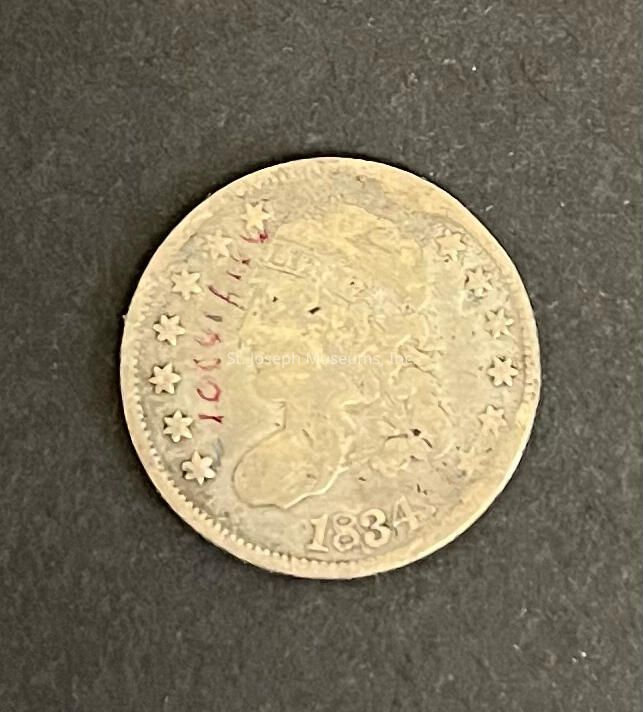 The image depicts an old coin with a design featuring a profile view of a person wearing a headband inscribed with the word “LIBERTY.” This coin, dated 1834 at the bottom, is surrounded by a series of stars along the edge. The coin appears worn, with visible signs of age and faint engraved details, contributing to its antique appearance. Additionally, there is red writing on the coin, although its significance is unclear. The background of the image is a textured, dark surface that contrasts with the coin’s lighter tone, highlighting its intricate detailing and historic character. The overall feel is one of historical curiosity, reflective of the period from which it originated.
