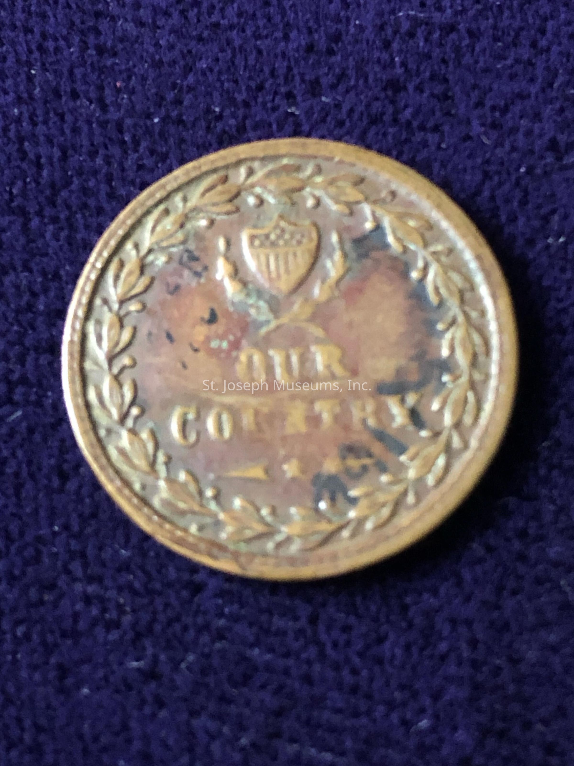 The image displays a vintage token or coin showcasing an embossed design. At its center, the text "OUR COUNTRY" is visible, surrounded by a decorative border featuring laurel leaves. Above the text, there is an emblem resembling a shield, potentially indicating a patriotic or national theme. The token has a tarnished brass or bronze hue, suggesting age and wear, which adds to its antique character. This circular token rests on a dark, textured fabric background, likely enhancing its historical and nostalgic aesthetic. The overall appearance suggests that the token might have been used for commemorative or collectible purposes.