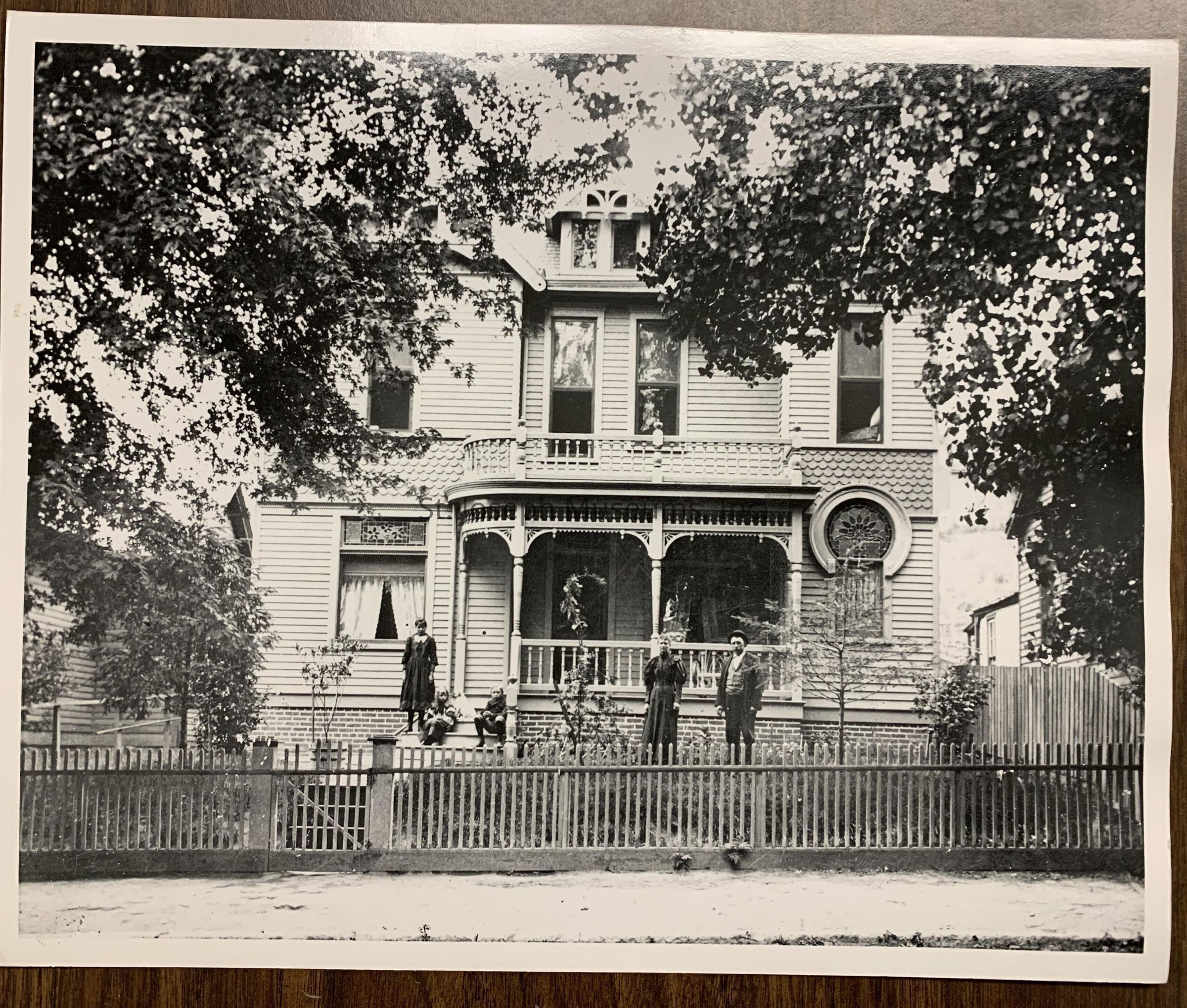 Black and white photograph showing a large, ornate Victorian-era house in St. Joseph, MIssouri surrounded by a wooden fence and lush trees, with several people, including a child, gathered in front of the structure. The house features decorative details like window trims and a circular stained glass window on the front facade. The trees provide partial shade, creating a dappled effect on the house's exterior. The people, dressed in period clothing, stand on the porch and near the gate.