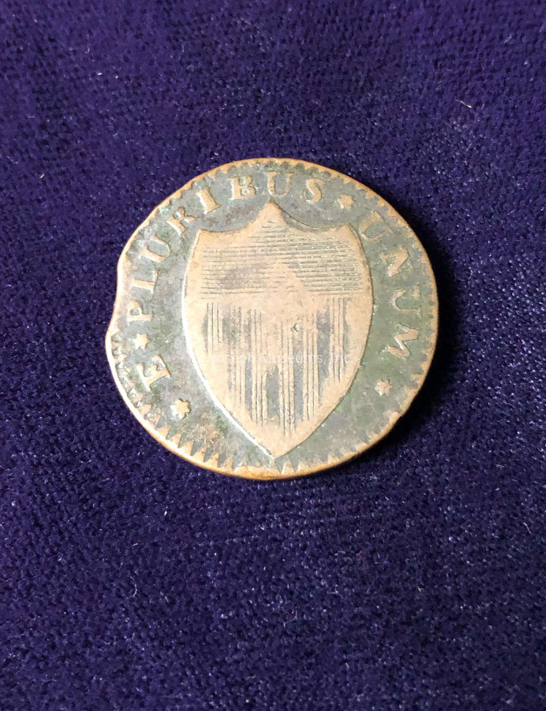 The coin features a prominent design with a large shield emblem at its center, flanked by a dotted rim. Surrounding the shield is an inscription spelling out "E PLURIBUS UNUM," which is a Latin phrase meaning "Out of many, one," encircling the top half of the coin. The shield itself has vertical stripes, adding to the intricate detailing of the coin's design. The overall color of the coin appears to be aged, with a patina that suggests it may be quite old or a collector's item. The coin's edge is slightly worn, consistent with its possible age.