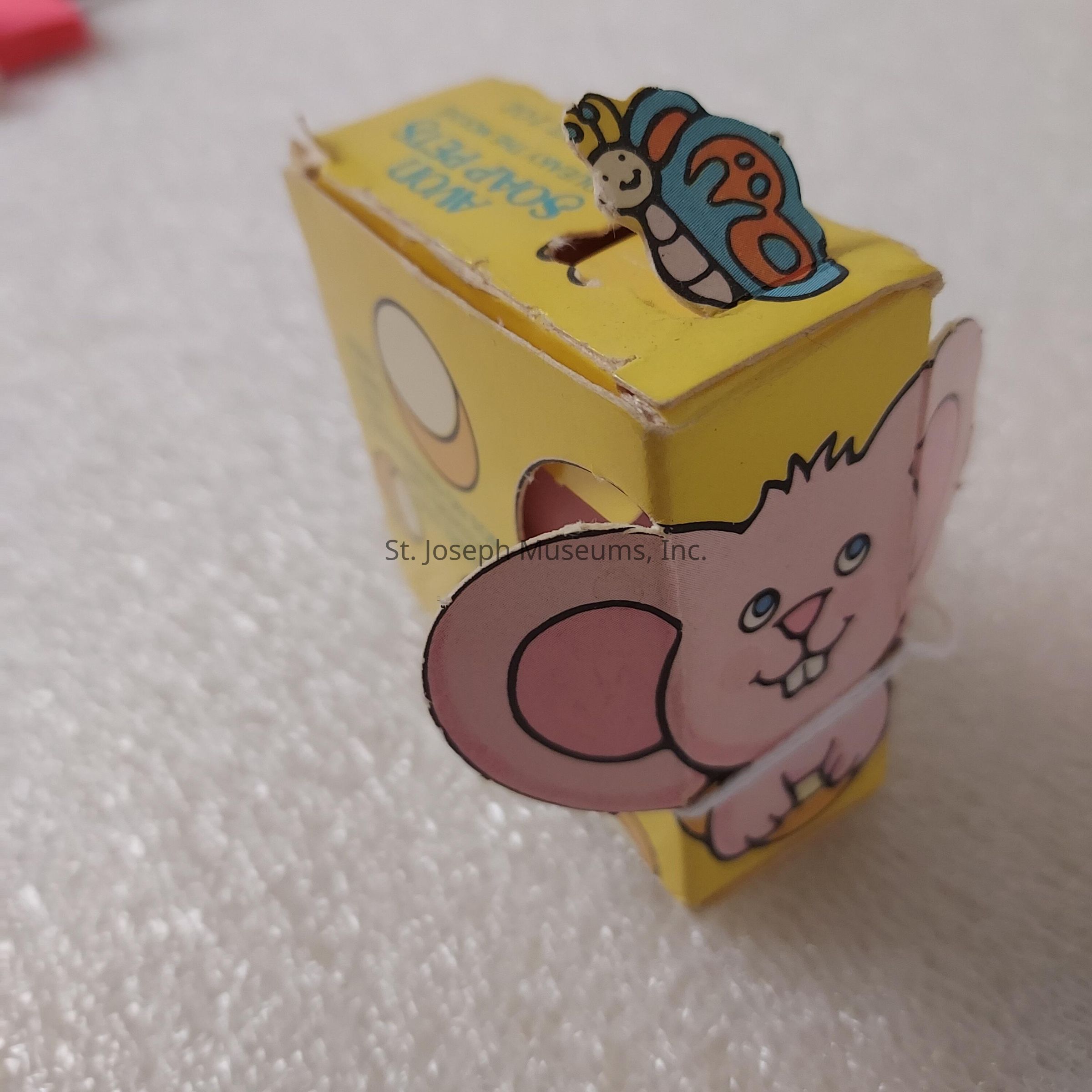The image displays a small, yellow cardboard box designed with playful illustrations. The box features the face and ears of a cartoon mouse, with a cheerful expression and big, pink ears set against the yellow background. On top of the box, there is a colorful butterfly illustration, adding a whimsical touch to the design. The box appears to be a product or toy package, likely for a children's item, as indicated by the vibrant colors and engaging animal graphics. The background is a textured, light-colored surface, providing a simple yet contrasting backdrop that highlights the box's vivid and lively appearance.