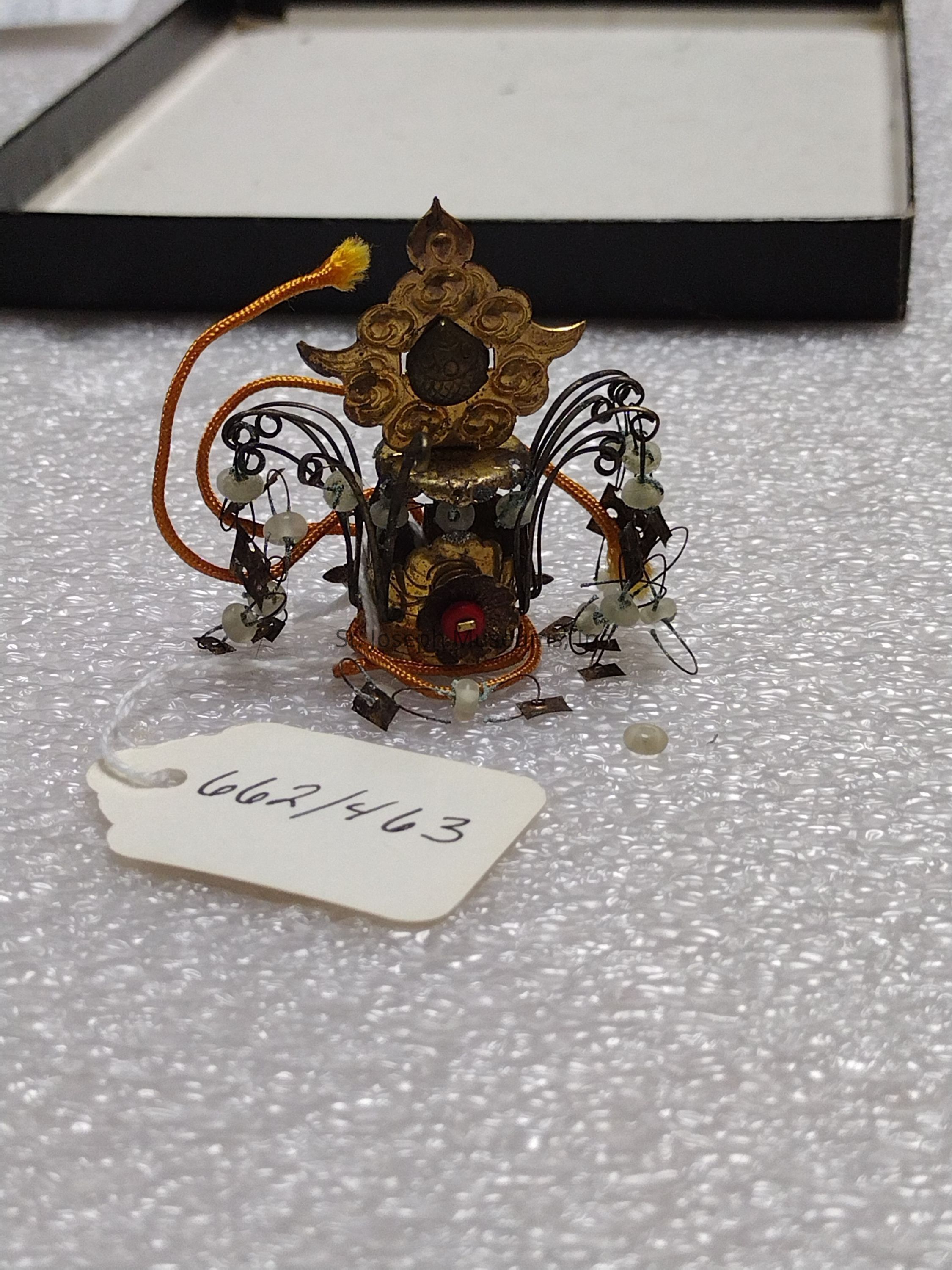 The image displays a small, intricate decorative object featuring an ornate design with a central cylindrical element adorned with a detailed golden motif at the top, reminiscent of a floral or flame-like structure, and surrounded by elegant curved metalwork that might suggest leaves or tendrils. Attached to these metal curvatures are small beads or ornaments, possibly made from jade or another precious material, enhancing the delicate craftsmanship. A bright orange cord loops around the sculpture, adding a splash of vibrant color. The piece is positioned on a textured white surface, possibly bubble wrap, suggesting it might be a fragile item intended for careful handling or storage. There is an accompanying white tag with the handwritten numbers "662/463" indicating it could be part of a collection or inventory, and behind this setup, there appears to be a partially open black box, further implying the object's potential purpose as a collectible or museum piece.