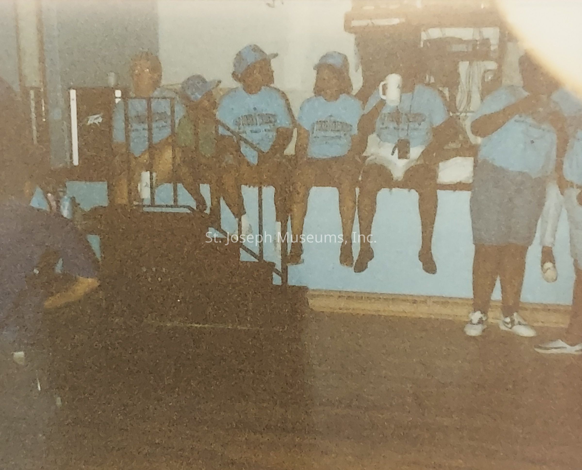 The image depicts a group of people sitting on a blue stage-like platform, wearing matching blue shirts and baseball caps, indicating they might be part of a team or club participating in a group event or gathering. Some individuals are holding cups, which suggests a casual social setting. The group spans both adults and children, and they appear relaxed and engaged in conversation. There is a wooden floor in front of them, and various pieces of equipment are visible in the background, possibly suggesting a recreational or community event taking place indoors. The overall mood seems friendly and informal.