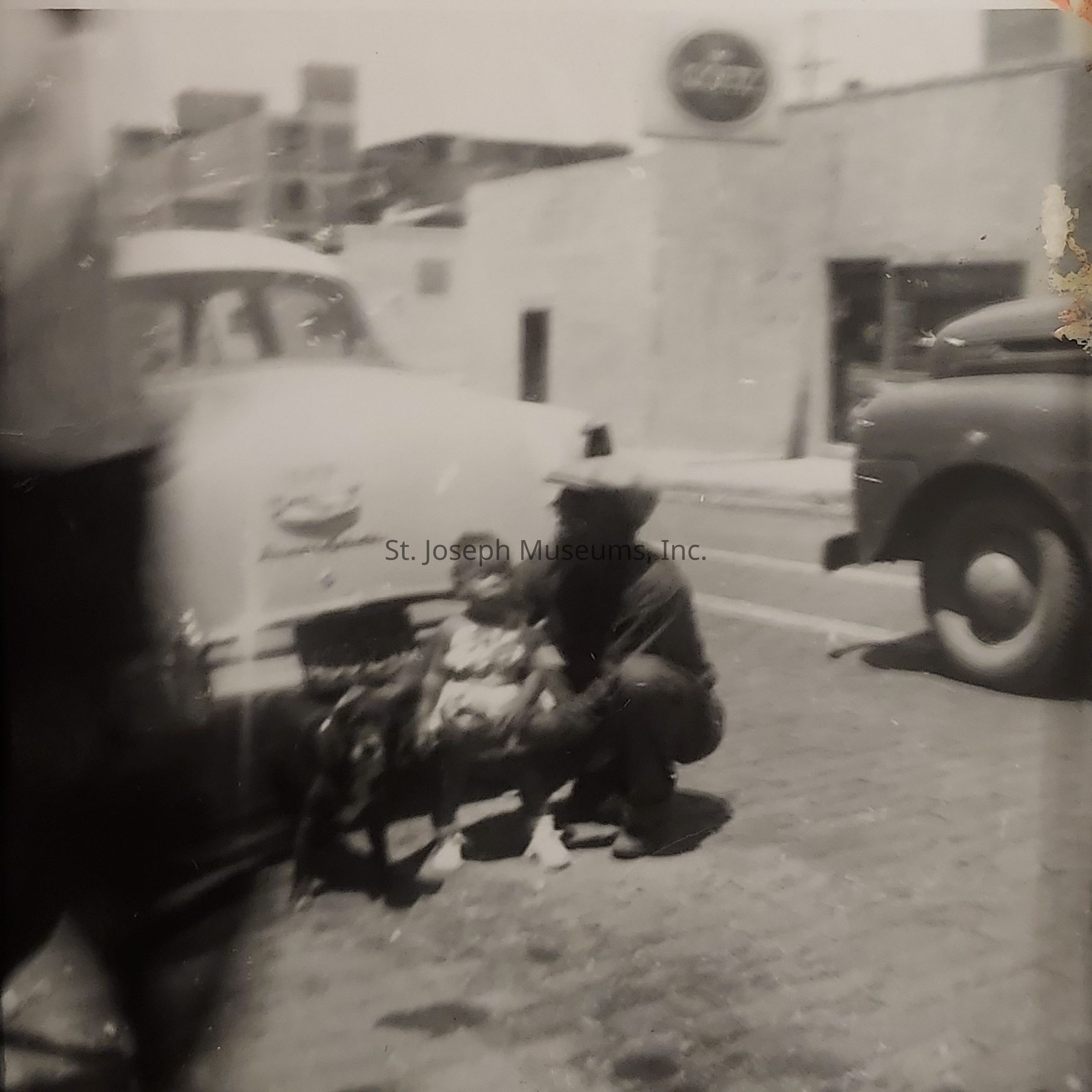 The black-and-white image depicts an outdoor scene where a young child, possibly sitting on the lap of an adult, is seen near the rear end of a parked vintage car, indicating an older time period. The adult, who appears to be squatting or kneeling, wears a hat and holds the child gently. Next to them, a dog is also present, seemingly leashed and calm. The background showcases an urban setting with a concrete building and another car parked on the street, suggesting a city environment. A sign with some text is visible above the building, hinting at a possible storefront or commercial area. The image, slightly blurred and marked with age, conveys a nostalgic atmosphere, capturing a candid moment in time where the focus lies on the relationship between the people and their pet amidst a bustling city backdrop. The simplicity and spontaneity of the composition evoke a sense of warmth and familial bonding.