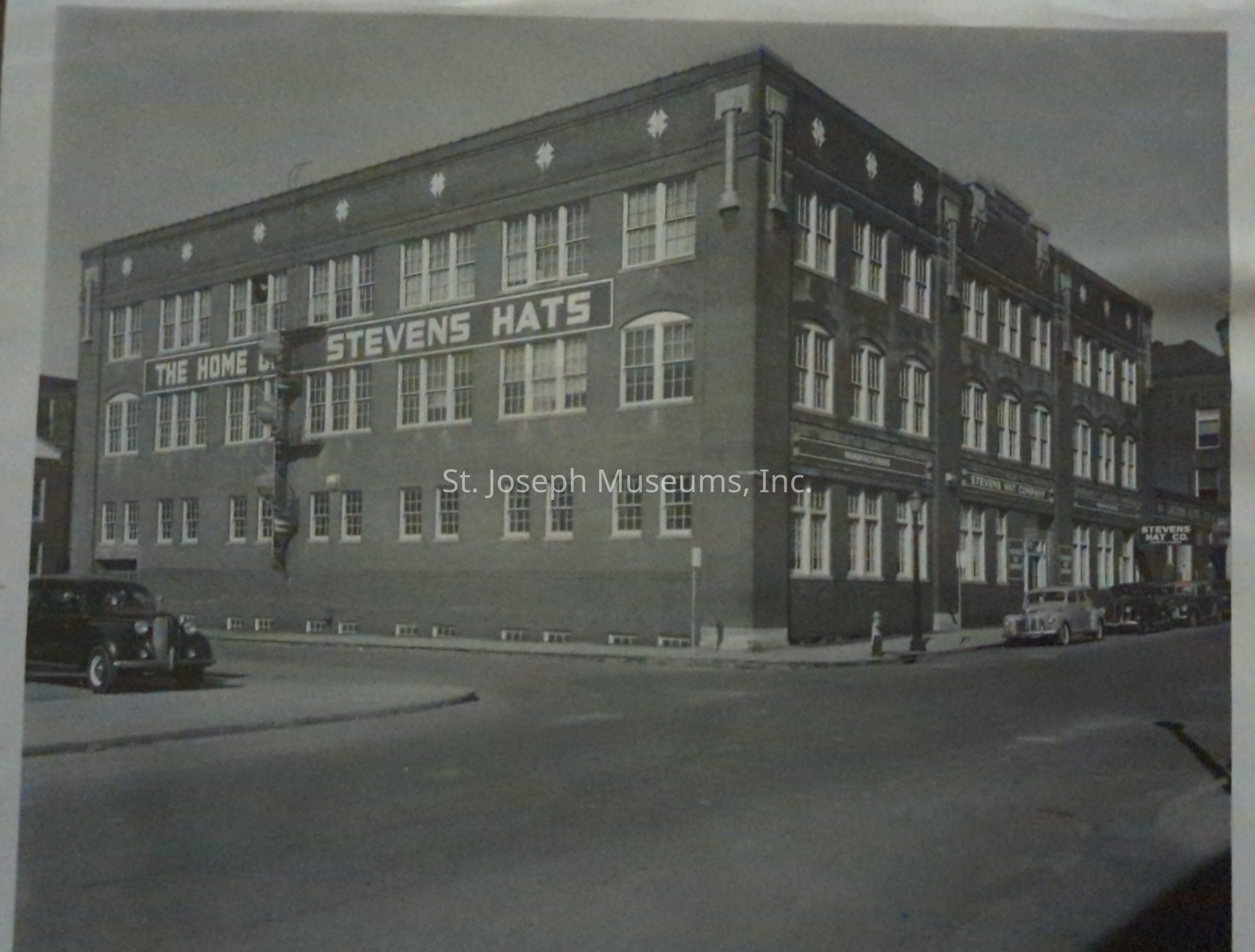 Black-and-white image depicting a historic multi-story brick building located at 314 South 7th Street, prominently featuring signage for "The Home of Stevens Hats." Classic cars are parked along the street, enhancing the vintage atmosphere of the scene. The architecture includes numerous windows and decorative cornices.
