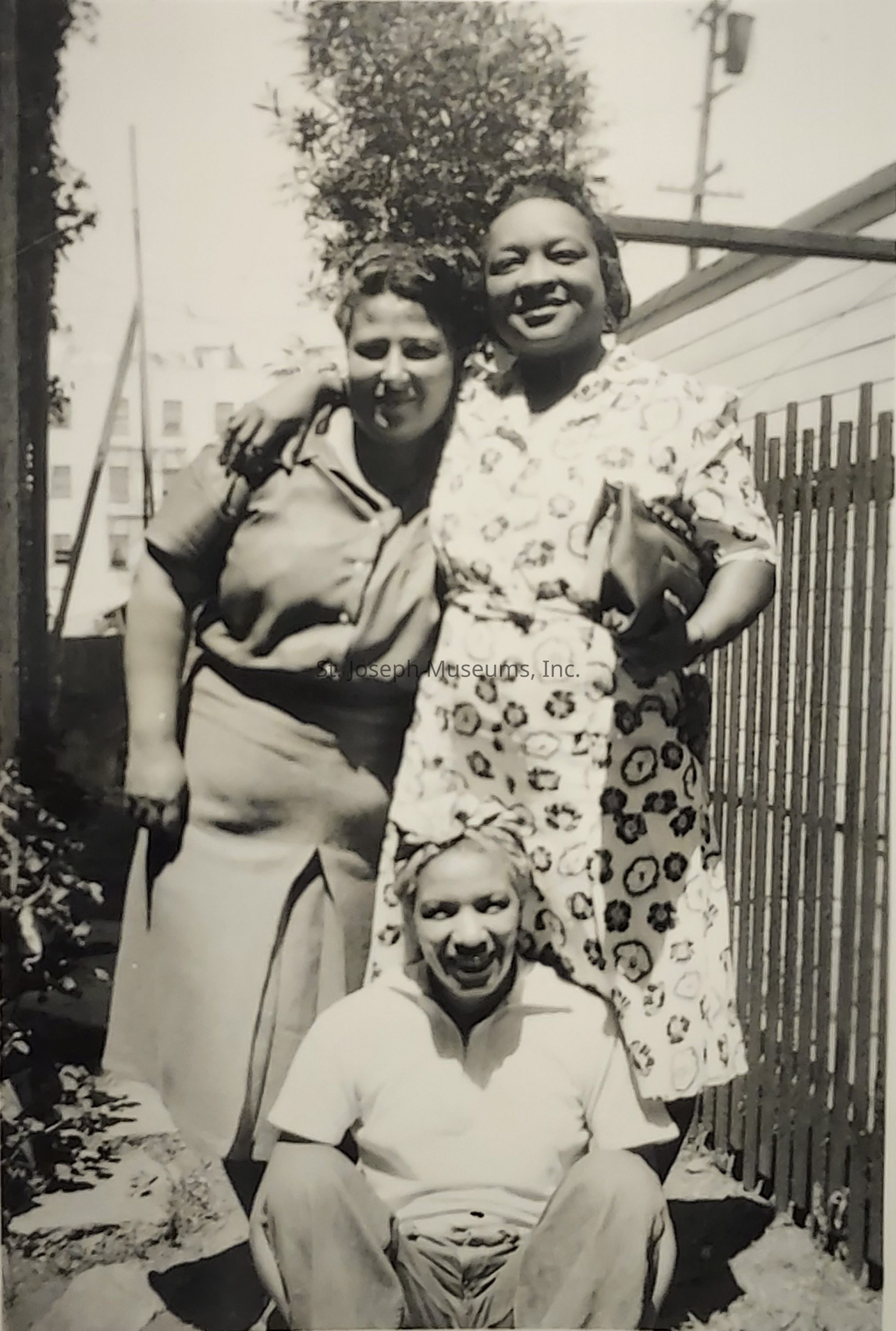The image features three individuals, two women and one seated person, posing together outdoors. The two women, one wearing a light, patterned dress, and the other in darker, plain attire, have their arms affectionately around each other, conveying a sense of camaraderie and warmth. The seated individual, positioned in front of them, is wearing a light-colored shirt and has a cheerful expression, enhancing the overall friendly and joyous mood of the image. The background features a fence, plants, and blurred buildings, suggesting an outdoor, perhaps urban setting. This black-and-white photograph captures a moment of togetherness, highlighting timeless themes of friendship and community.