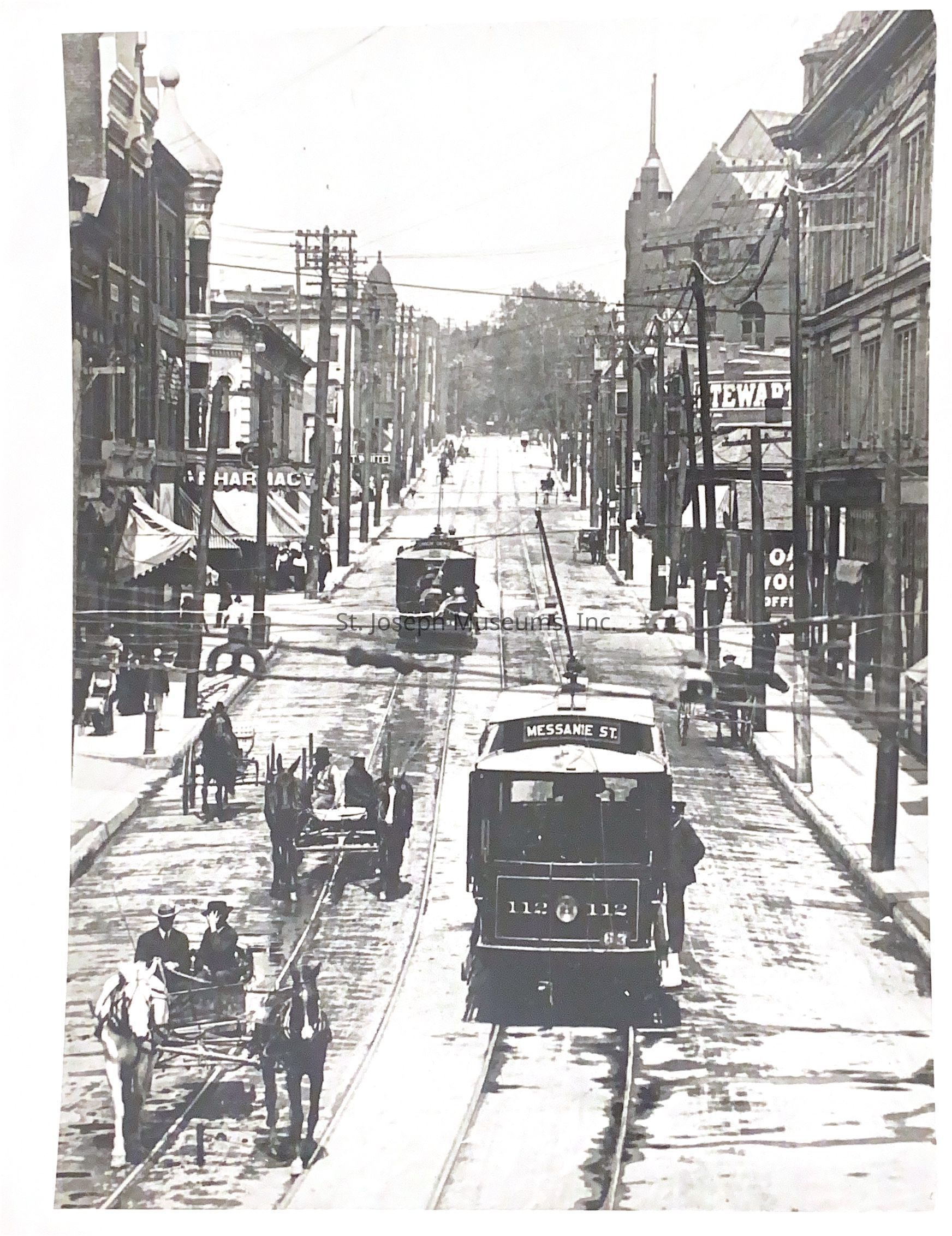 Black-and-white photograph showcasing a bustling street scene with a trolley car traveling along Frederick Avenue from 8th Street. Several horse-drawn carriages move alongside pedestrians on the sidewalks. Buildings line both sides, with visible storefronts, including a pharmacy. Overhead wires stretch across the street.