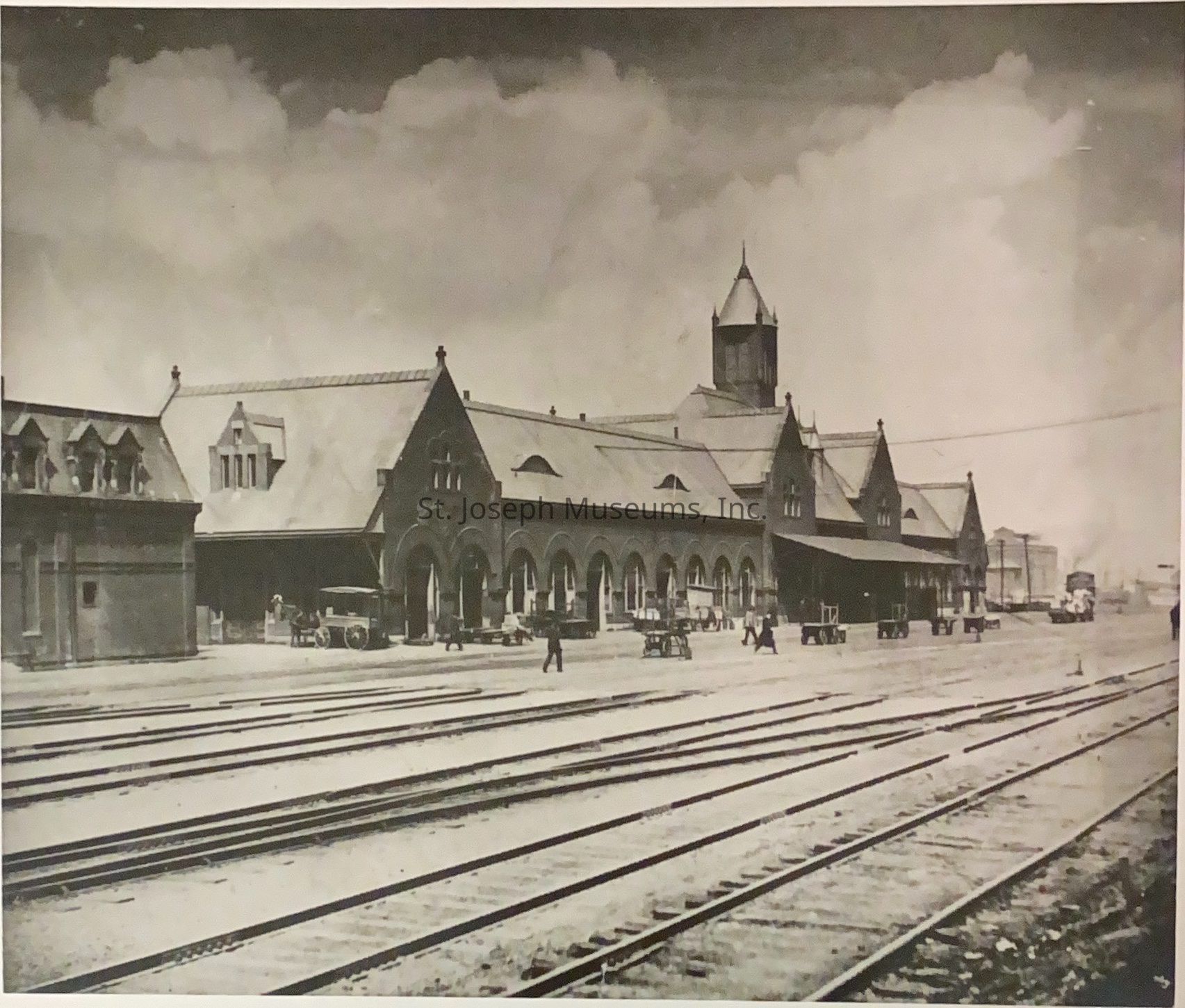 Black-and-white image depicting Union Station in Kansas City, Missouri, showcasing its historical architecture with a large, central building featuring pointed roofs and numerous arches. Railroad tracks stretch across the foreground, while several people and carts are visible near the station. The background contains a few structures and clouds.