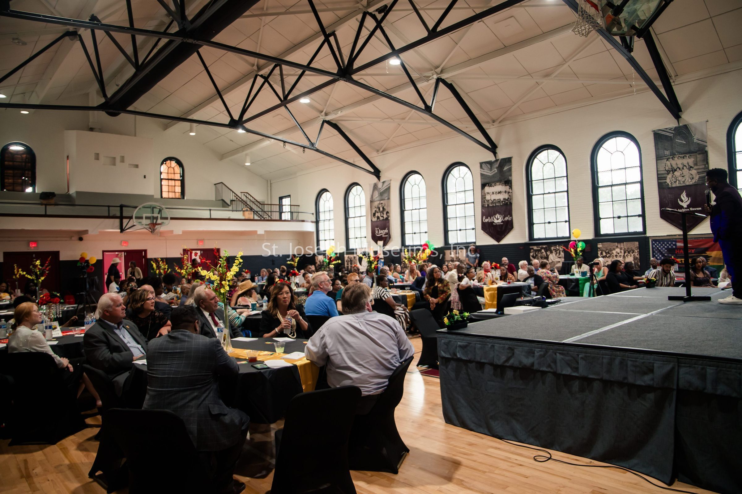 The image showcases a lively event taking place in a spacious gymnasium, where numerous attendees are seated at round tables, engaged in conversation and enjoying the gathering. The decor is elegant, featuring floral centerpieces on tables and festive balloons adding pops of color to the environment. The room is filled with natural light streaming through large arched windows along the walls, which are adorned with banners. In the foreground, a person stands on a stage with a microphone, indicating that speeches or performances may be part of the event, enhancing the lively and social atmosphere.
