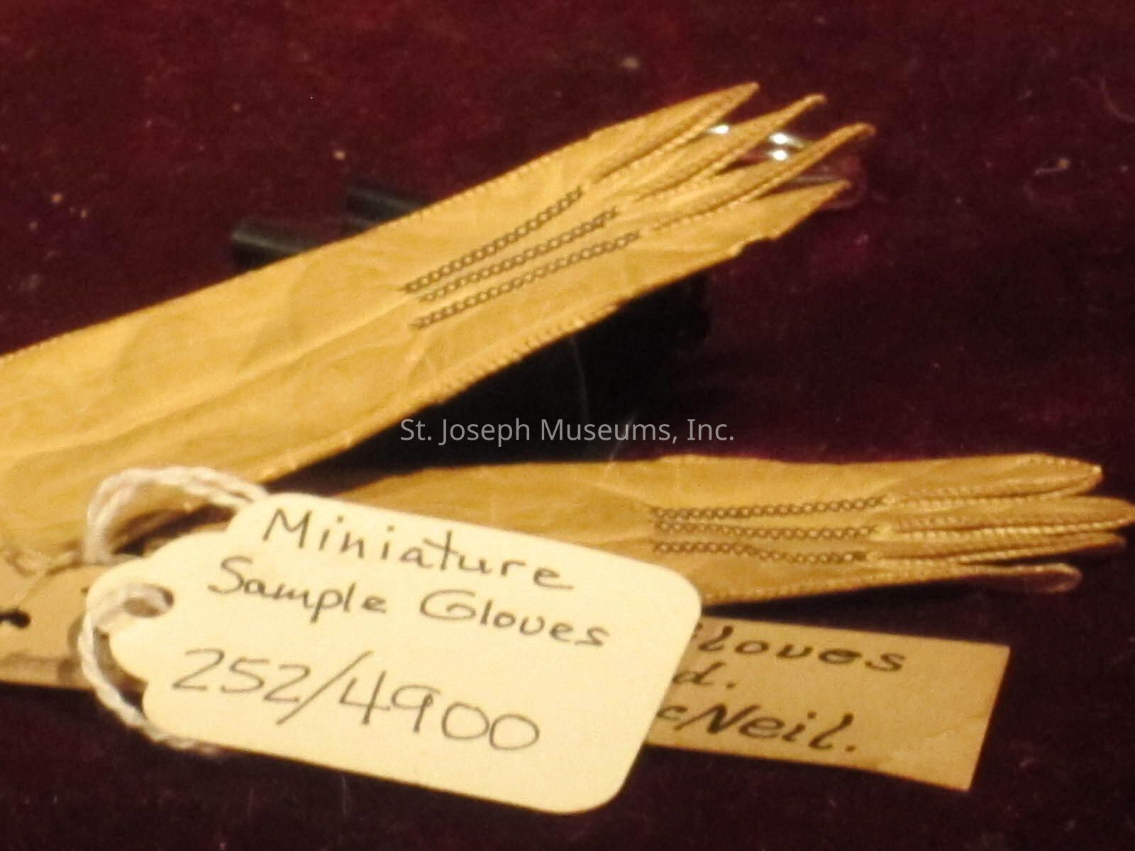 The image features a pair of miniature sample gloves with intricate stitching placed on a dark, possibly velvet, surface. Attached to the gloves is a label made of sturdy paper or card, tied with a string, with handwritten text that reads "Miniature Sample Gloves 252/4900," indicating they are part of a limited edition or collection. The gloves, appearing to be made of a light brown material, possibly leather, are finely crafted with detailed stitching along the fingers and back. The background consists of a dark red or maroon fabric, providing a rich contrast to the light-colored gloves. A partially visible additional note is placed underneath, suggesting further descriptive or cataloging information, indicating the importance or value of these gloves, possibly for a museum artifact collection or a special exhibit piece. The entire composition captures a sense of historical significance or specialty in fashion, highlighting craftsmanship and attention to detail, especially in miniature collectible items, making them unique and noteworthy in the context presented.