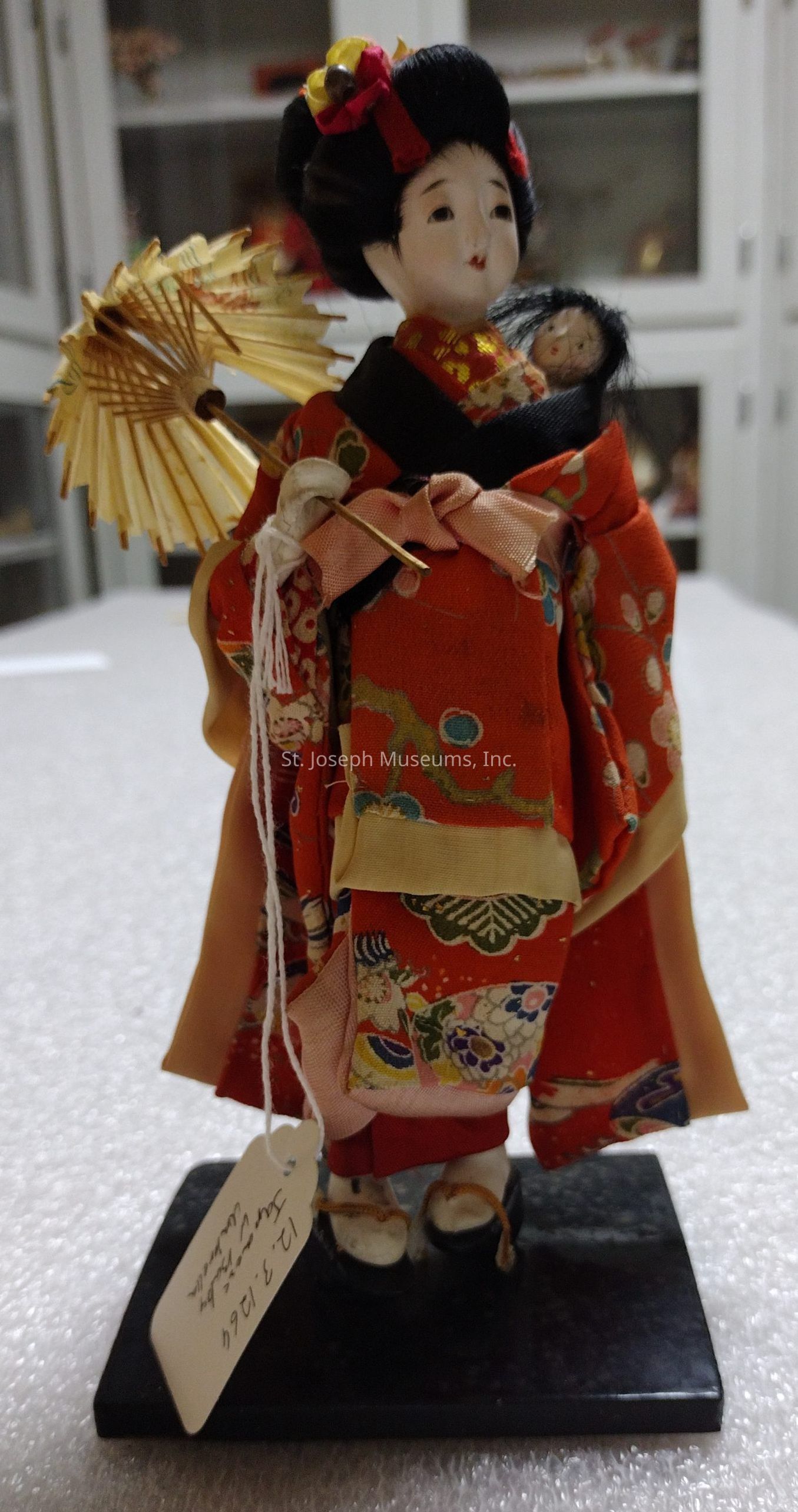 The image depicts a traditional Japanese doll dressed in an intricately patterned kimono adorned with vibrant floral designs in shades of red, green, and beige. The doll is holding a small, delicate parasol in one hand, which complements the elegant hairstyle adorned with a colorful ribbon and hair accessory. Nestled in the folds of the kimono is a smaller doll, adding a layer of charm and detail to the figure's overall presentation. The doll stands on a polished black base, and a tag is attached with a string, suggesting it may be part of a collection or exhibit. The backdrop shows a softly blurred interior setting, likely a display case or shelves, emphasizing the doll as the focal point.