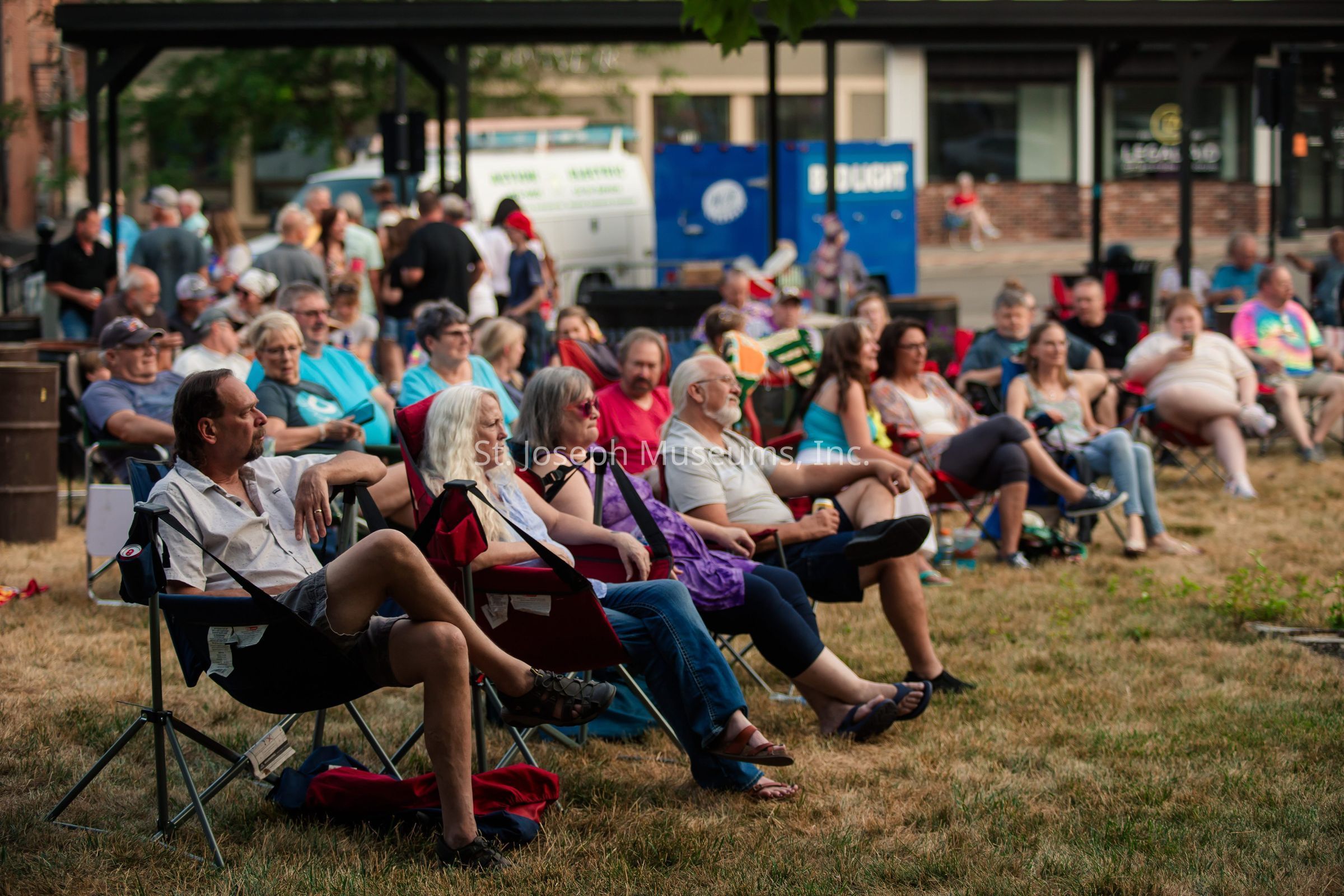 In the image, a diverse group of people is seated in an outdoor setting on a grassy area, sitting in various types of foldable and camping chairs, appearing to enjoy a public event or gathering. The attendees are casually dressed, suggesting a relaxed and informal atmosphere. Some are holding drinks, while others are simply sitting comfortably and engaging in conversation or watching something that the setting might offer. Visible in the background are other individuals and groups standing and mingling, indicating a social and communal vibe to the scene. The backdrop includes some urban elements such as a building with windows, a vehicle, and a Bud Light sign, suggesting this event is taking place in a city environment. The warm, natural lighting implies the event could be occurring in the late afternoon or early evening, providing a welcoming environment for attendees to relax and socialize in a communal setting. This gathering likely serves as a pleasant opportunity for members to connect, converse, or enjoy entertainment, reflecting a sense of community and leisure.