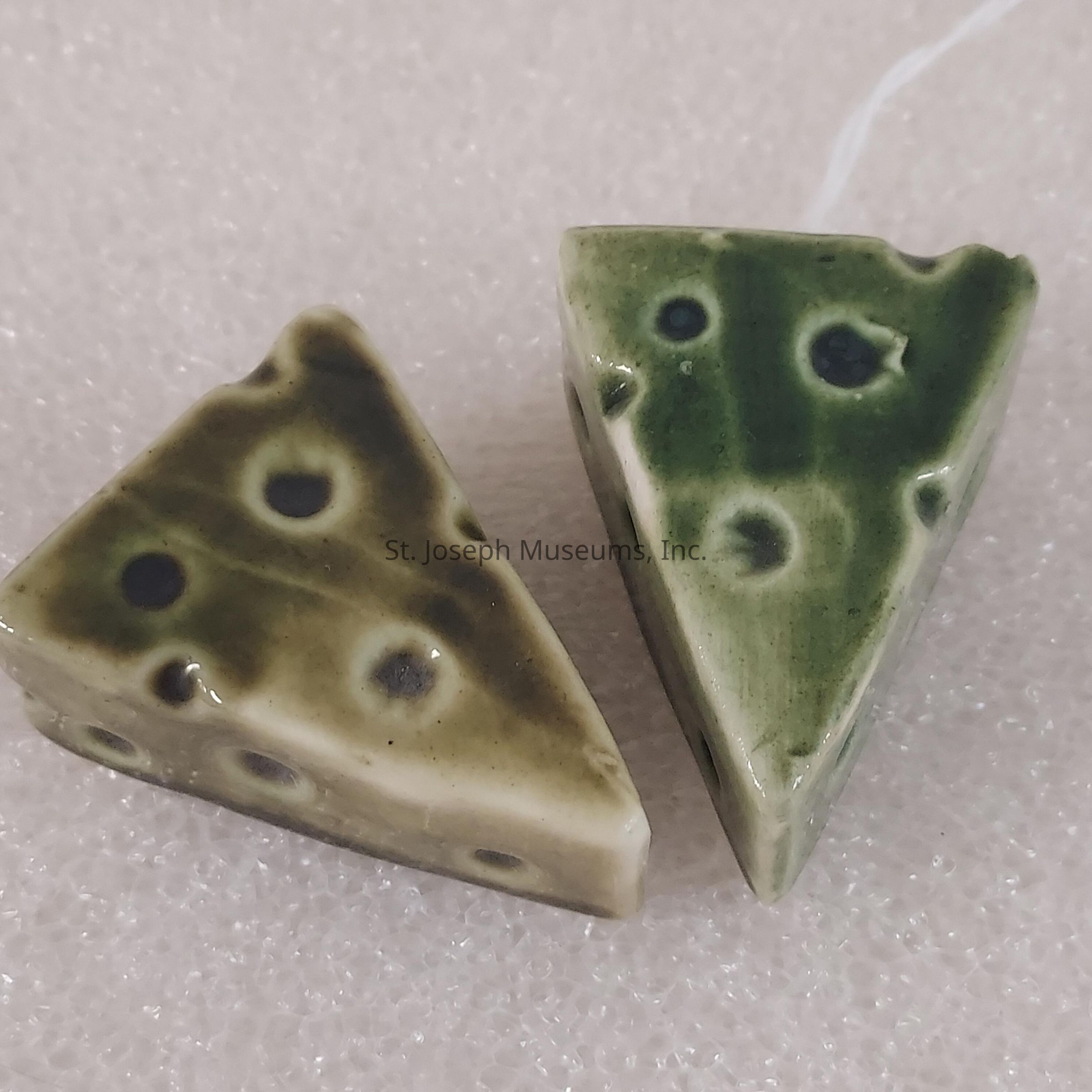 The image showcases two ceramic items shaped like slices of cheese on a textured surface. Both pieces are triangular, mimicking the appearance of classic cheese wedges with a series of irregular, scattered holes that suggest the aged Swiss variety. The left piece is distinguished by its earthy brown color, with darker brown spots creating a realistic look akin to a rustic, aged cheese. The right piece, in contrast, is shaded in olive green with dark green spots, offering a more whimsical take on the cheese wedge concept. Each piece is glazed, lending them a glossy finish that enhances their visual appeal. The surface they rest on appears to be a light, speckled material, possibly foam or fabric, providing a gentle backdrop that contrasts with the ceramic pieces. These artistic renditions of cheese evoke both humor and craftsmanship, blending familiar food imagery with the creative expression often found in ceramic art. The simplicity and charm make them interesting decor items or conversation pieces, showcasing the creator's attention to detail in rendering familiar textures and forms.