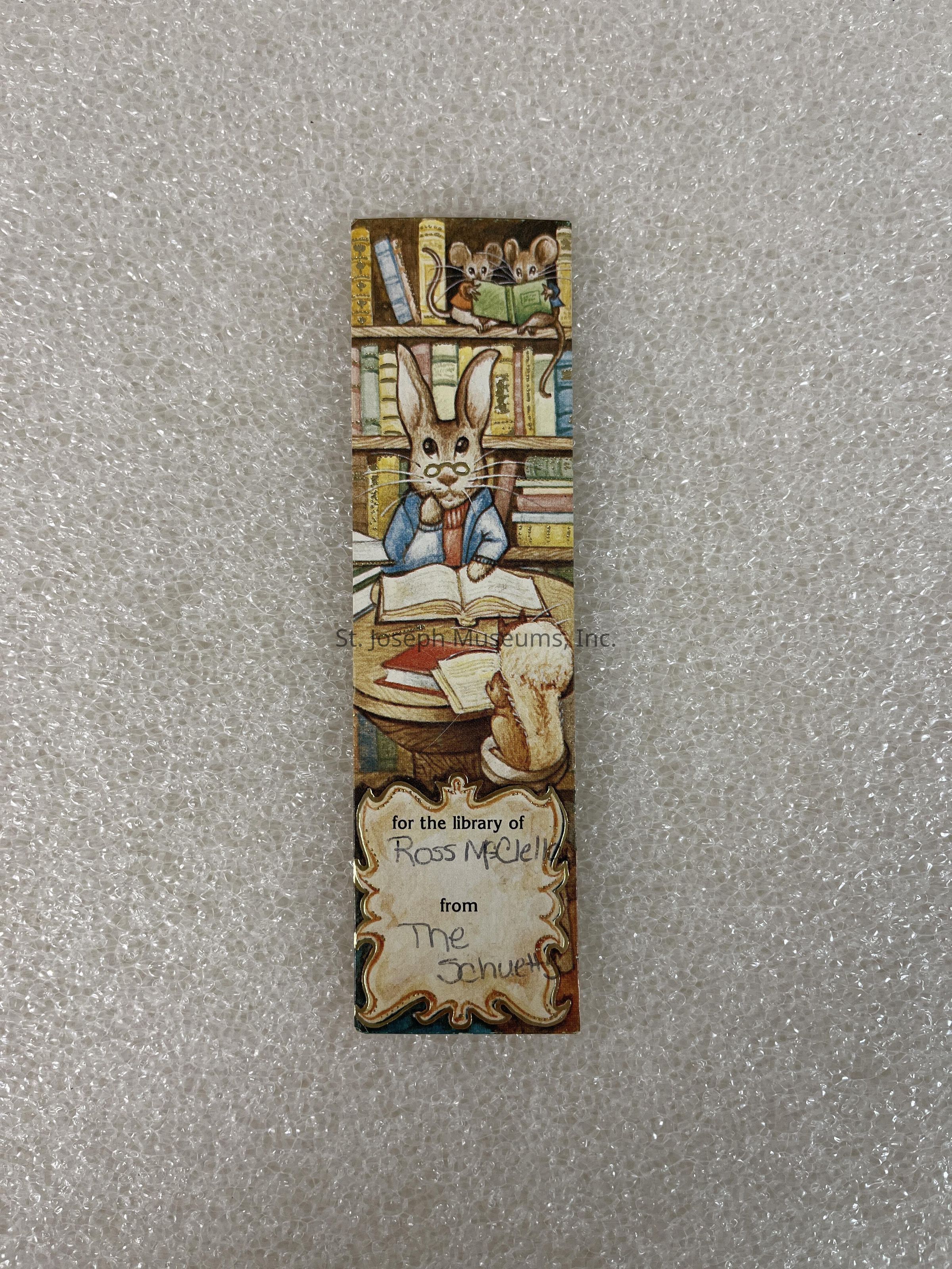 The image depicts a whimsical bookmark featuring illustrations of anthropomorphic animals in a library setting. At the center, a rabbit, wearing glasses and a blue jacket, is engrossed in reading a book on a desk surrounded by more books and papers. Above, two mice are perched on bookshelves filled with various colored books, and one mouse is reading a small green book. At the bottom, there is a decorative area with handwritten text that reads, "for the library of Ross McDell from The Schwett," suggesting the bookmark was a personal gift. The entire scene is set against a backdrop of a textured, light surface that contrasts with the detailed, colorful design of the bookmark.
